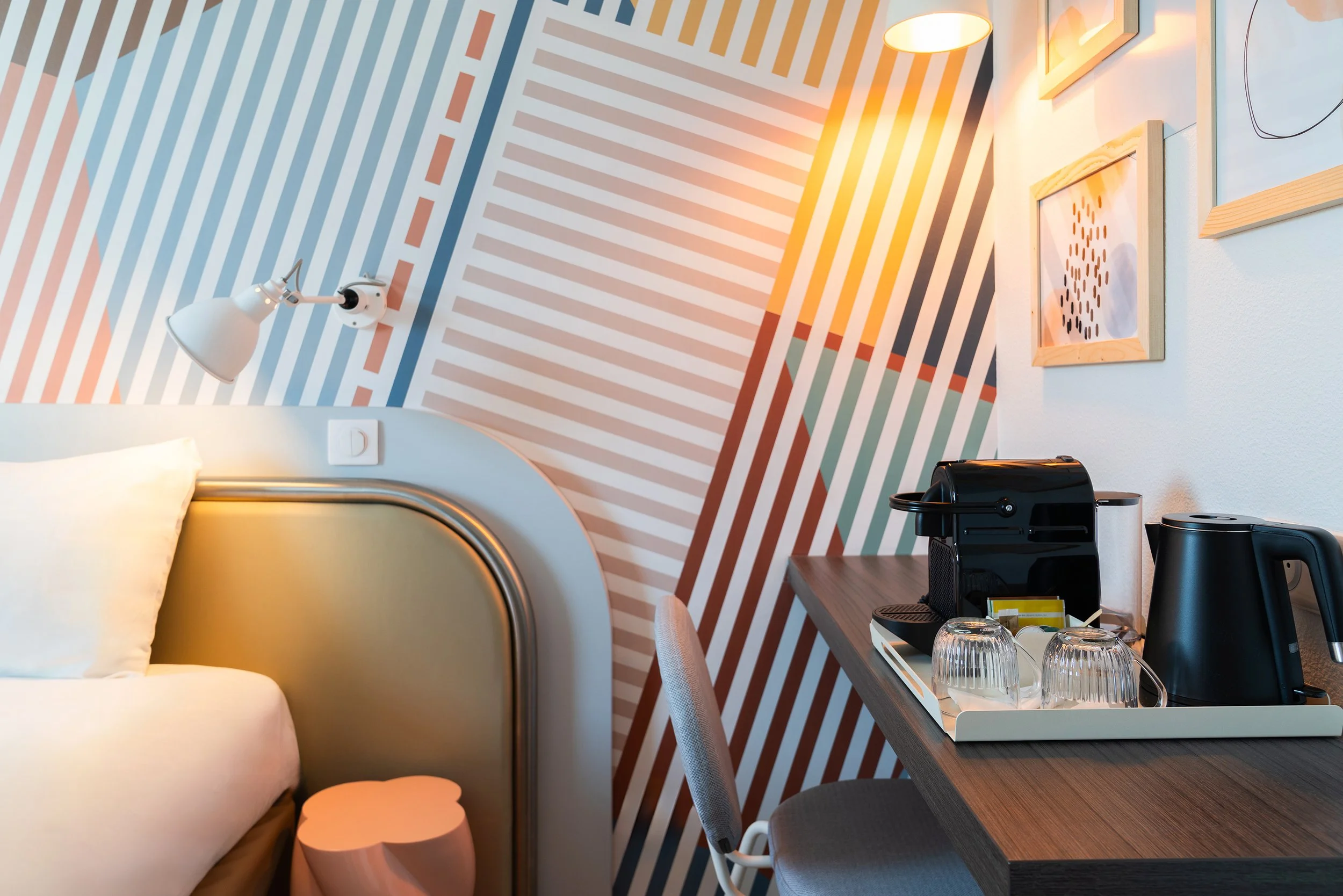 Intérieur d'une chambre d'hôtel avec un lit, une lampe, un mur décoratif avec des rayures colorées, un bureau avec une machine à café, une bouilloire, et des verres