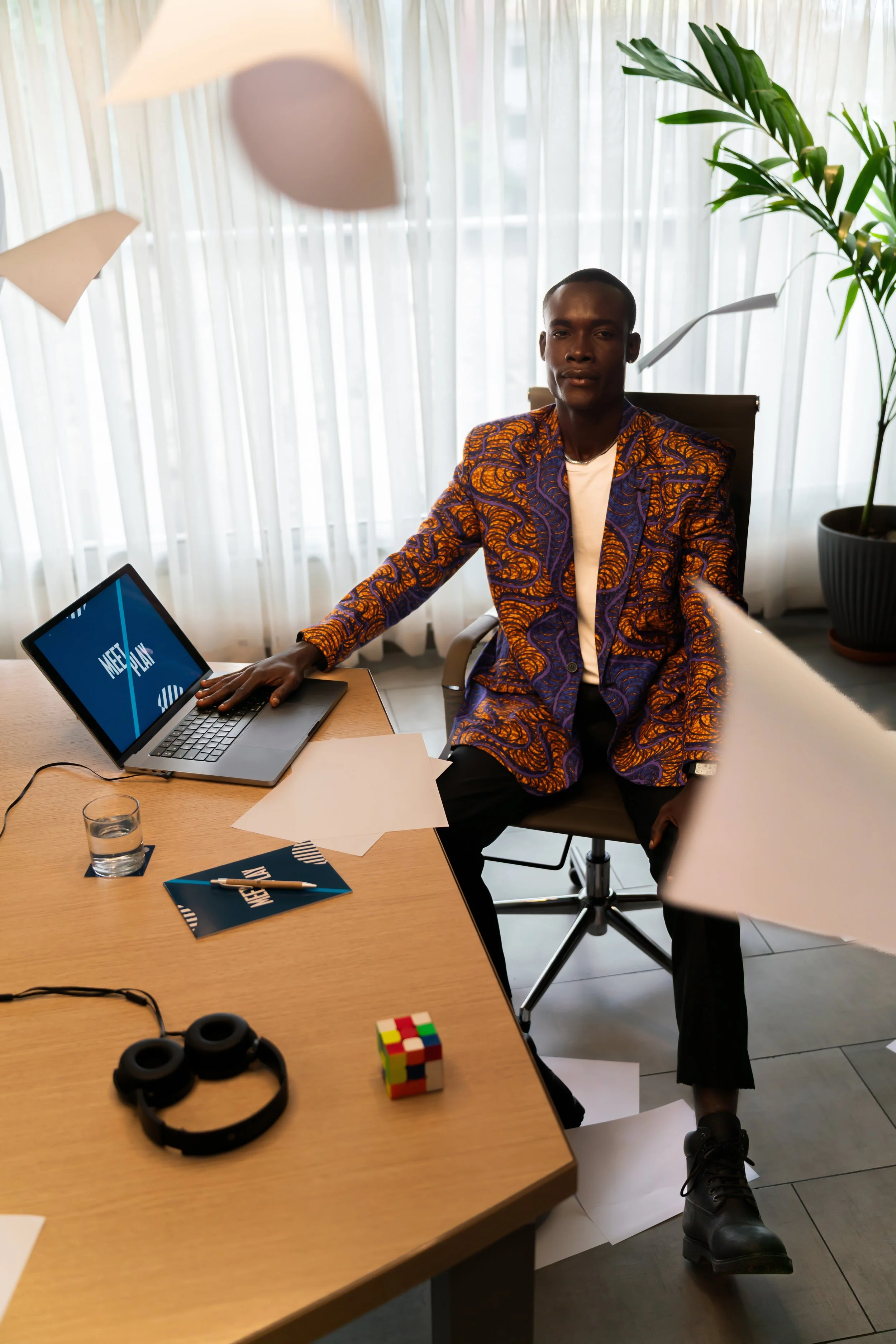 Homme assis à un bureau en costume coloré, entouré de papiers, d'un ordinateur portable affichant 'Meet & Play', d'un verre d'eau, d'un casques audio, dans un bureau lumineux avec des rideaux blancs et une grande plante.