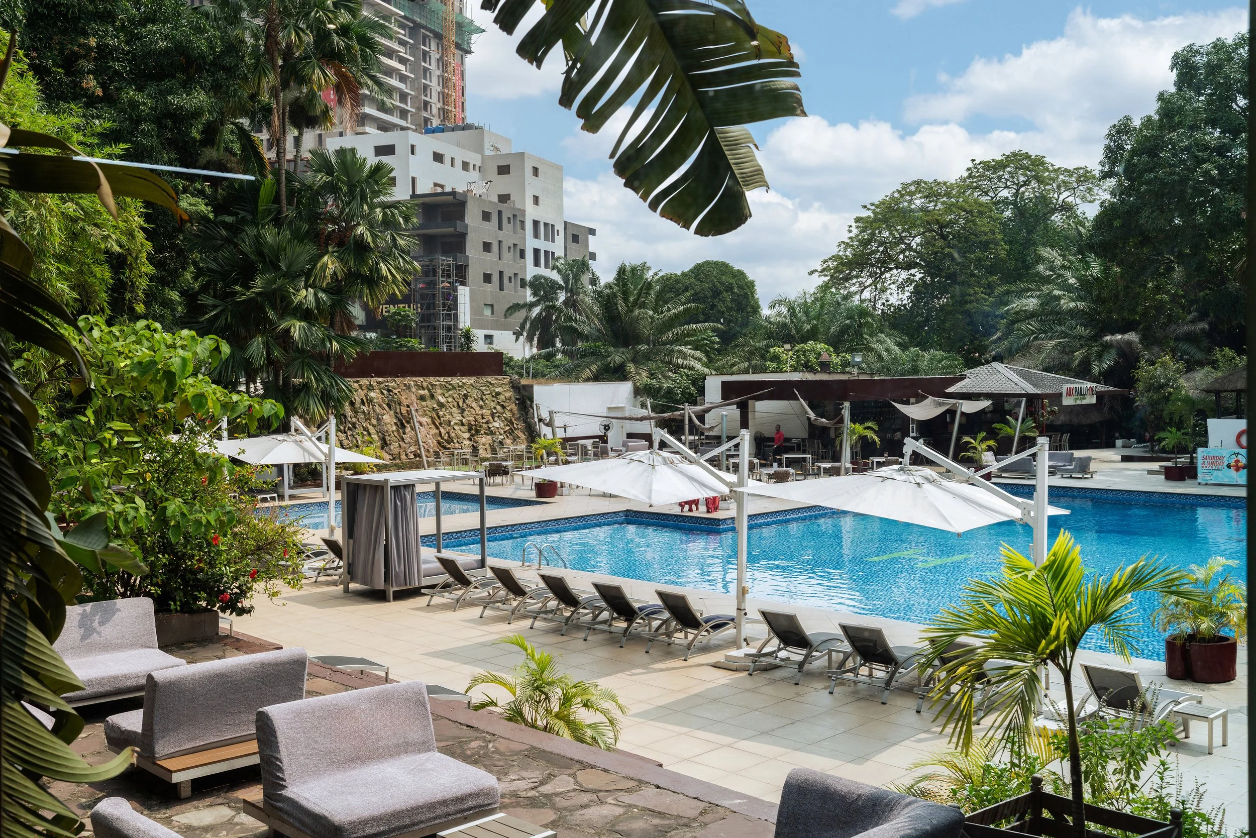 Piscine extérieure avec des chaises longues, parasols blancs, et végétation luxuriante dans un environnement urbain avec des bâtiments modernes en arrière-plan.
