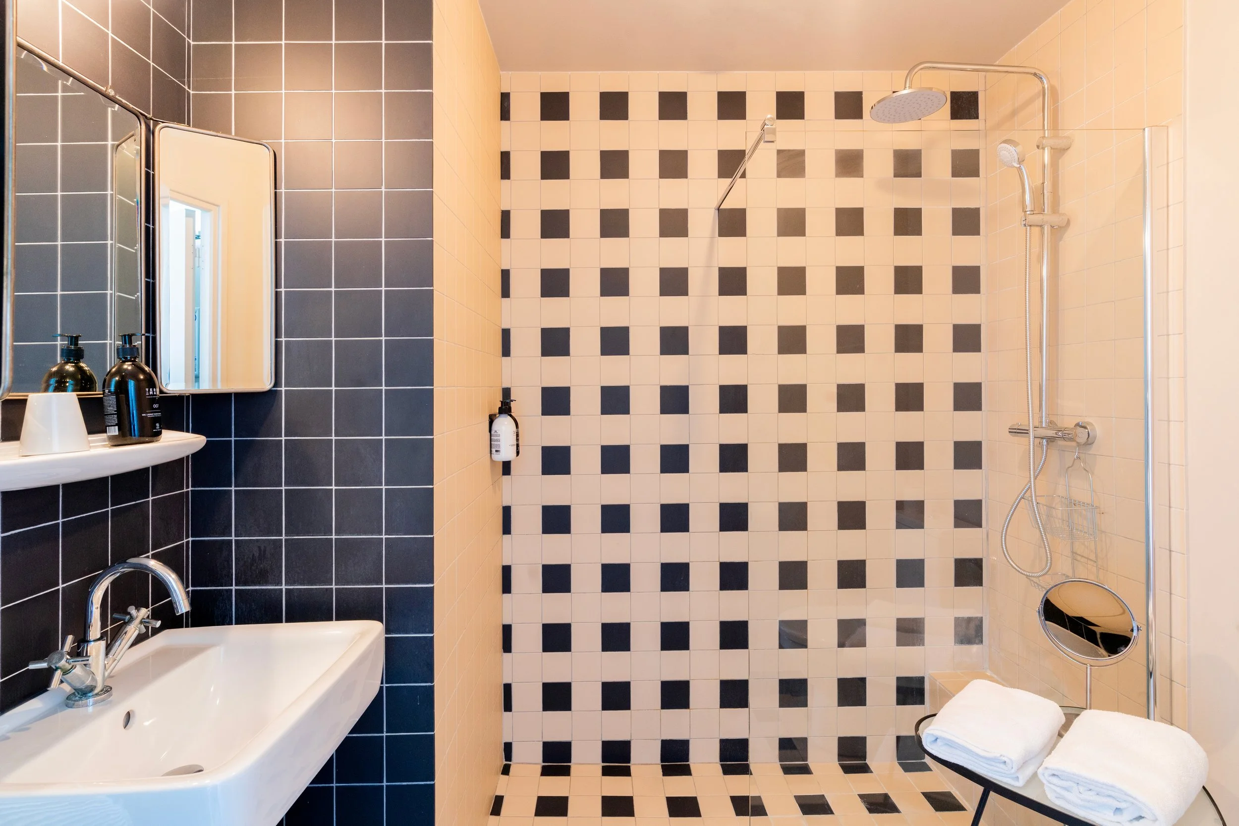 Salle de bain moderne avec un mur noir et blanc à carreaux, une cabine de douche en mosaïque, un lavabo blanc, des miroirs, des bouteilles de savon, et des serviettes blanches pliées sur une petite table.