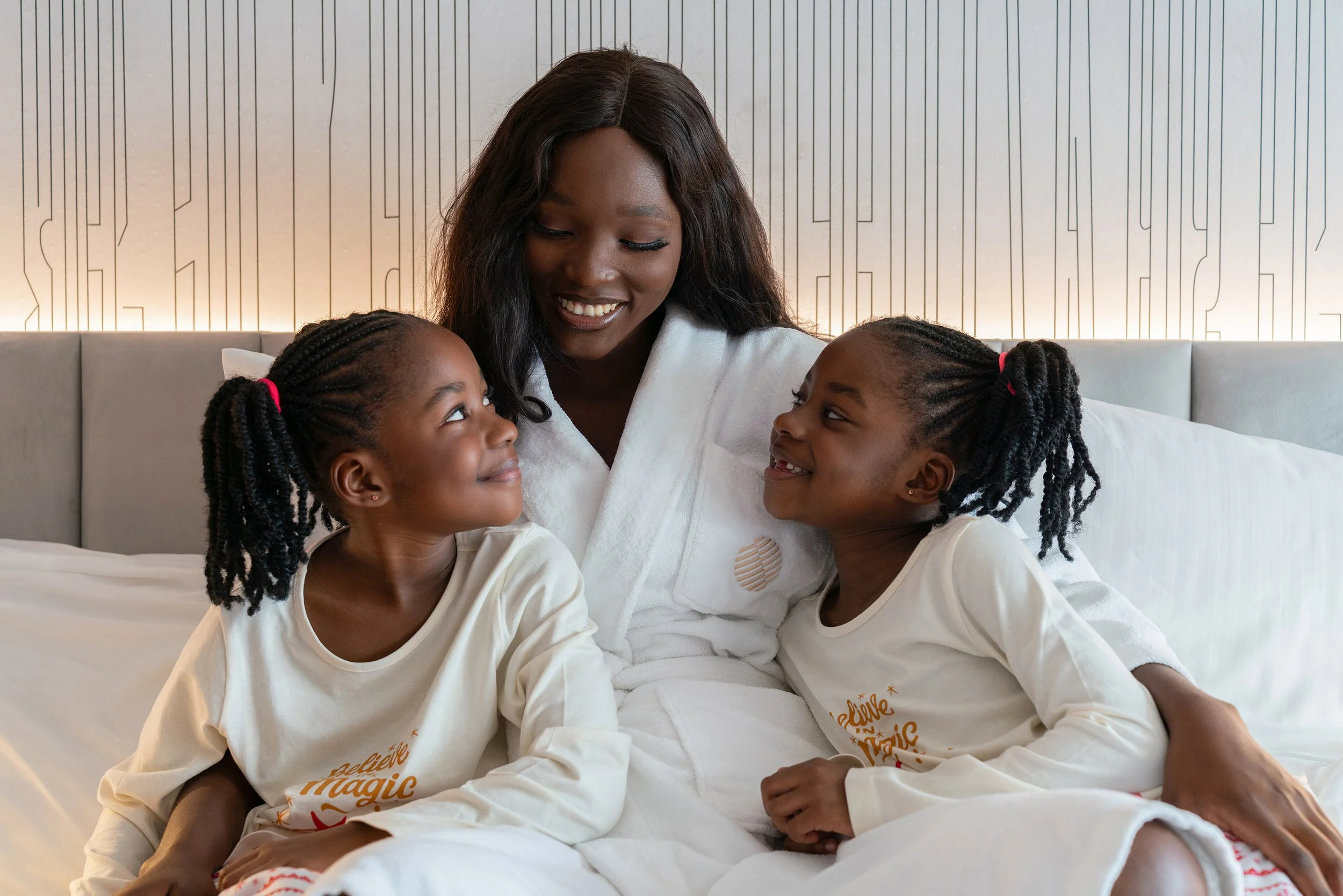 Une mère et ses deux jeunes filles souriantes assises sur un lit dans une chambre moderne.
