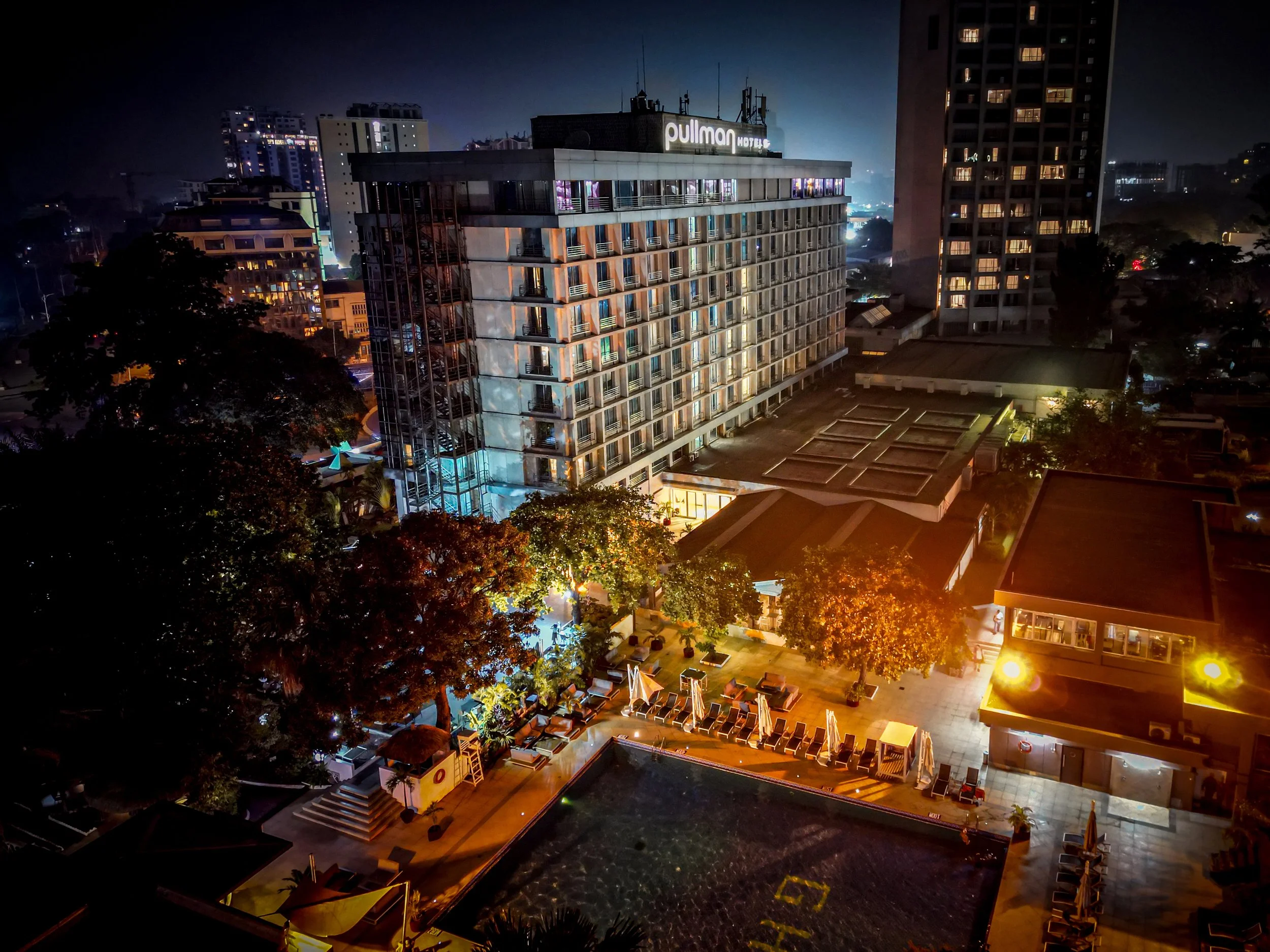Vue nocturne aérienne d'un hôtel urbain avec piscine, arbres, chaises longues et bâtiments modernes illuminés.