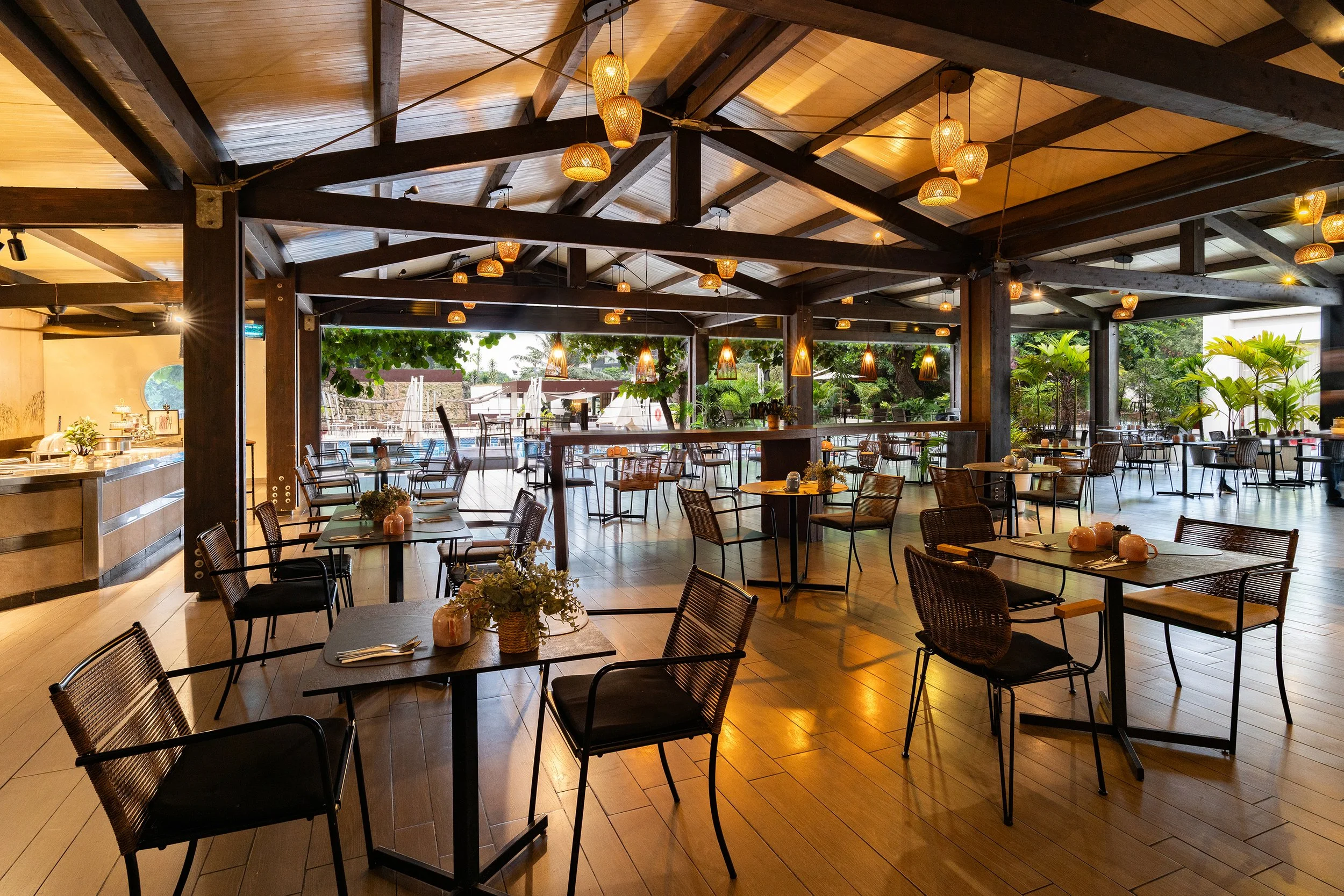Salle d'un restaurant avec décoration en bois, tables avec napperons et vases de fleurs, éclairage tamisé, et espace extérieur avec vue sur une piscine.