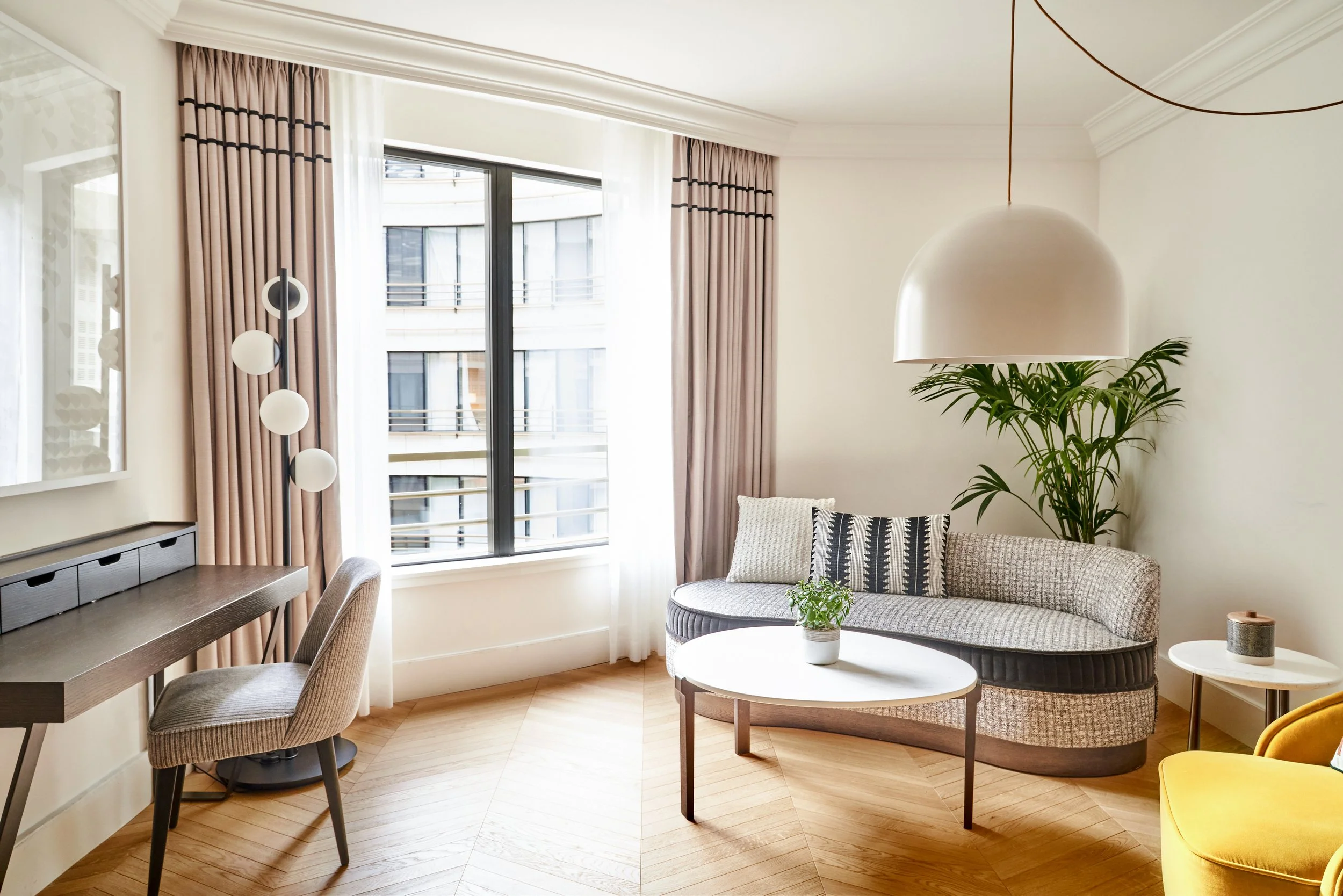 Salon moderne avec grande fenêtre, canapé en tissus avec coussin, plante verte, lampe suspendue blanche, table basse ronde, bureau avec chaise, décoration minimaliste.
