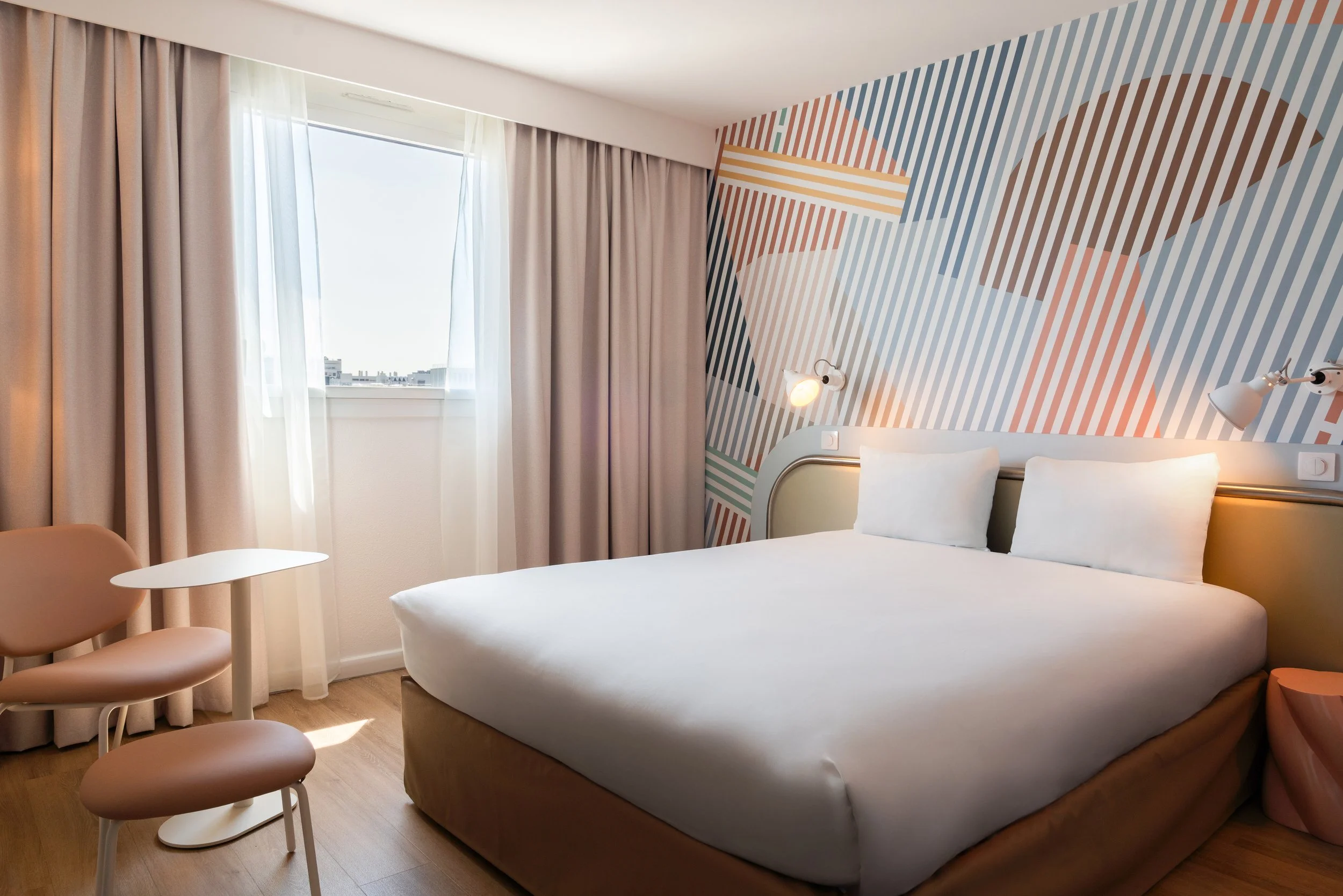Chambre d'hôtel moderne avec lit double, mur avec motif géométrique coloré, fenêtres avec rideaux beiges, table et deux chaises beige.