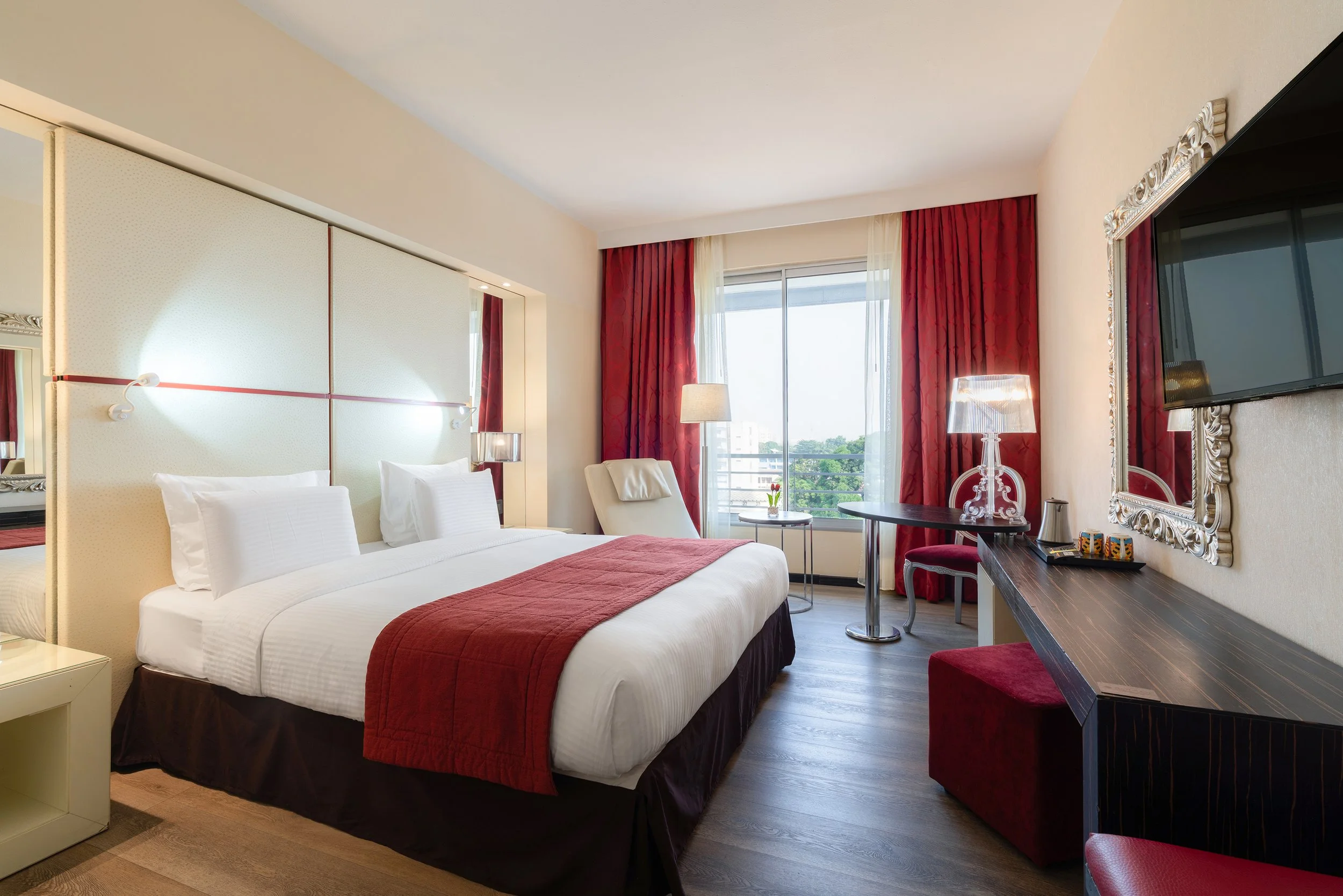 Chambre d'hôtel avec lit double, rideaux rouges, fauteuil, bureau en bois, télévision murale et grande fenêtre avec vue sur l'extérieur.