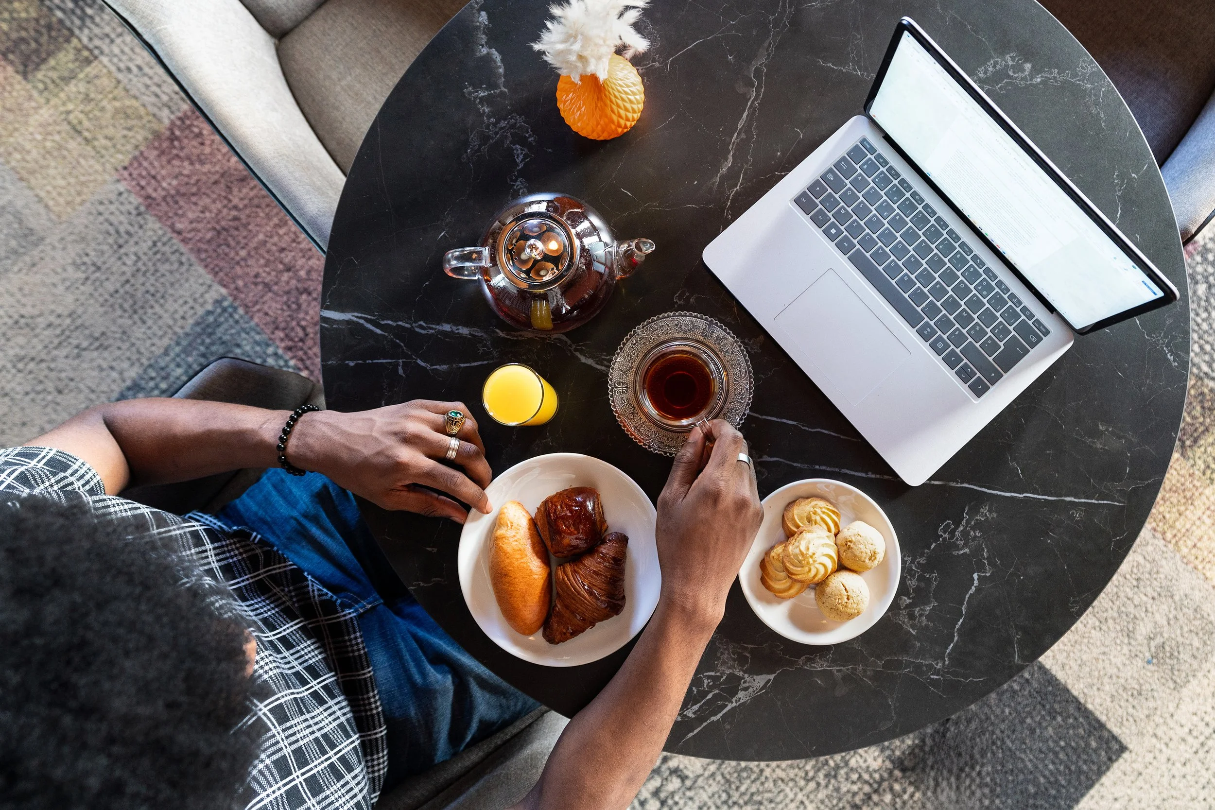 Une personne prend un café avec des croissants, un pain, et des biscuits, devant un ordinateur portable sur une table en marbre noir, avec une théière, un verre de jus d'orange, et un verre de thé, dans un café.