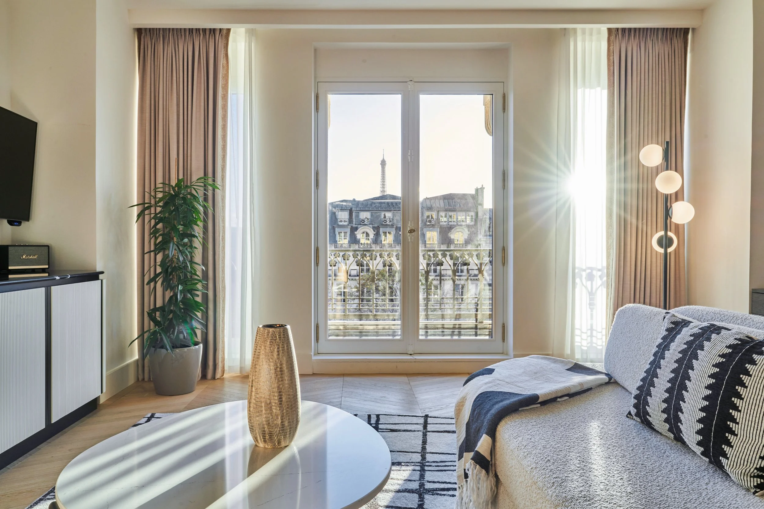 Salon lumineux avec grande fenêtre donnant sur la tour Eiffel, canapé beige avec coussins noirs et blancs, table basse ronde avec vase en porcelaine, lampe surmontée de trois globes, rideaux beiges, et une grande plante verte à gauche.