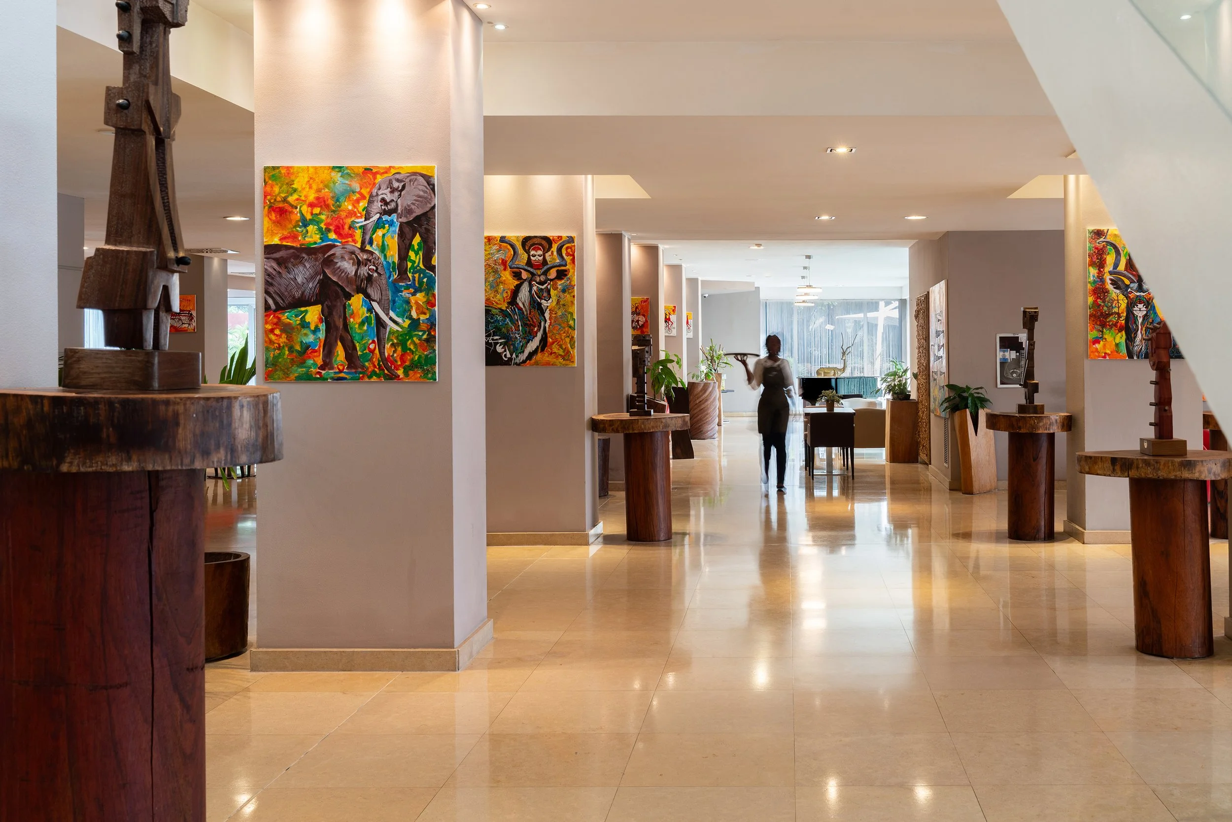 Intérieur d'un hôtel ou d'une galerie d'art avec des tableaux colorés d'éléphants et d'oiseaux, plusieurs sculptures en bois, des plantes vertes, et une serveuse qui se déplace