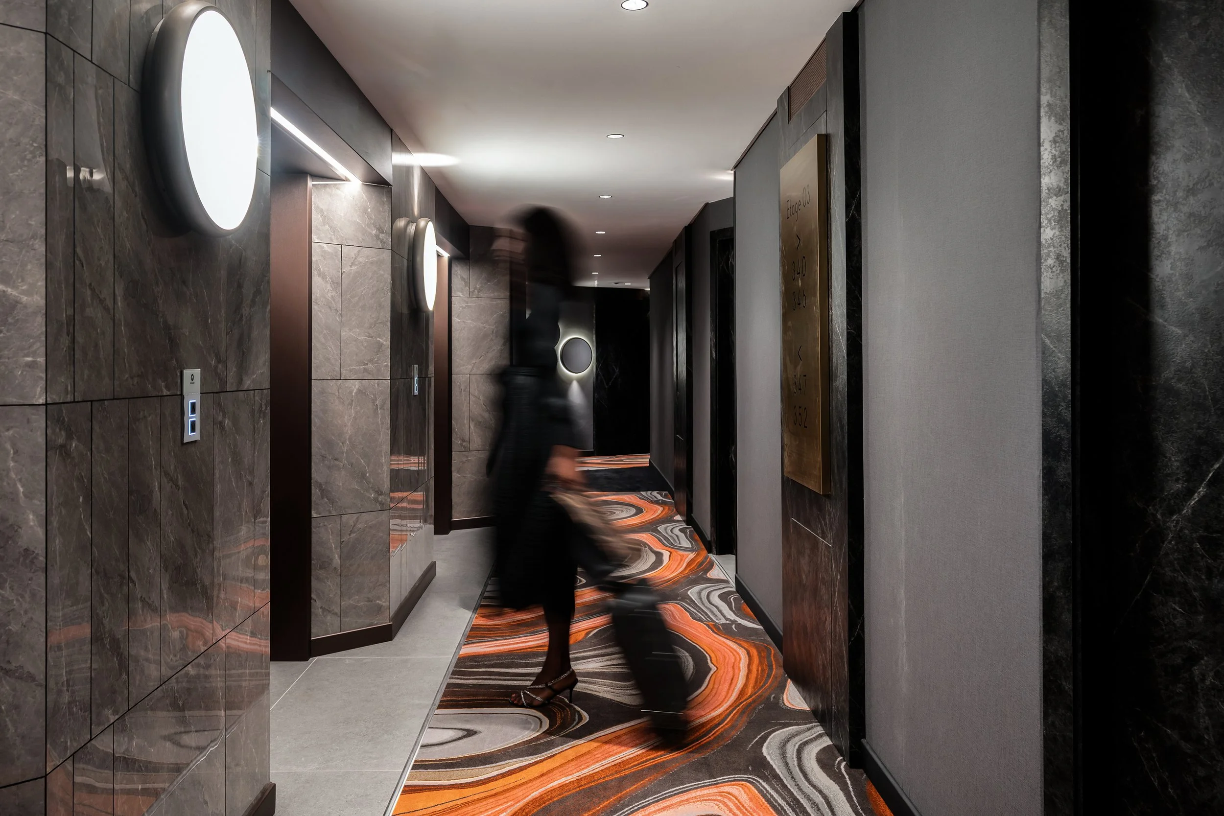 Couloir d'hôtel avec murs en marbre et tapis décoratif aux motifs orange, gris et noir, personne marchant en portant un sac à roulettes.