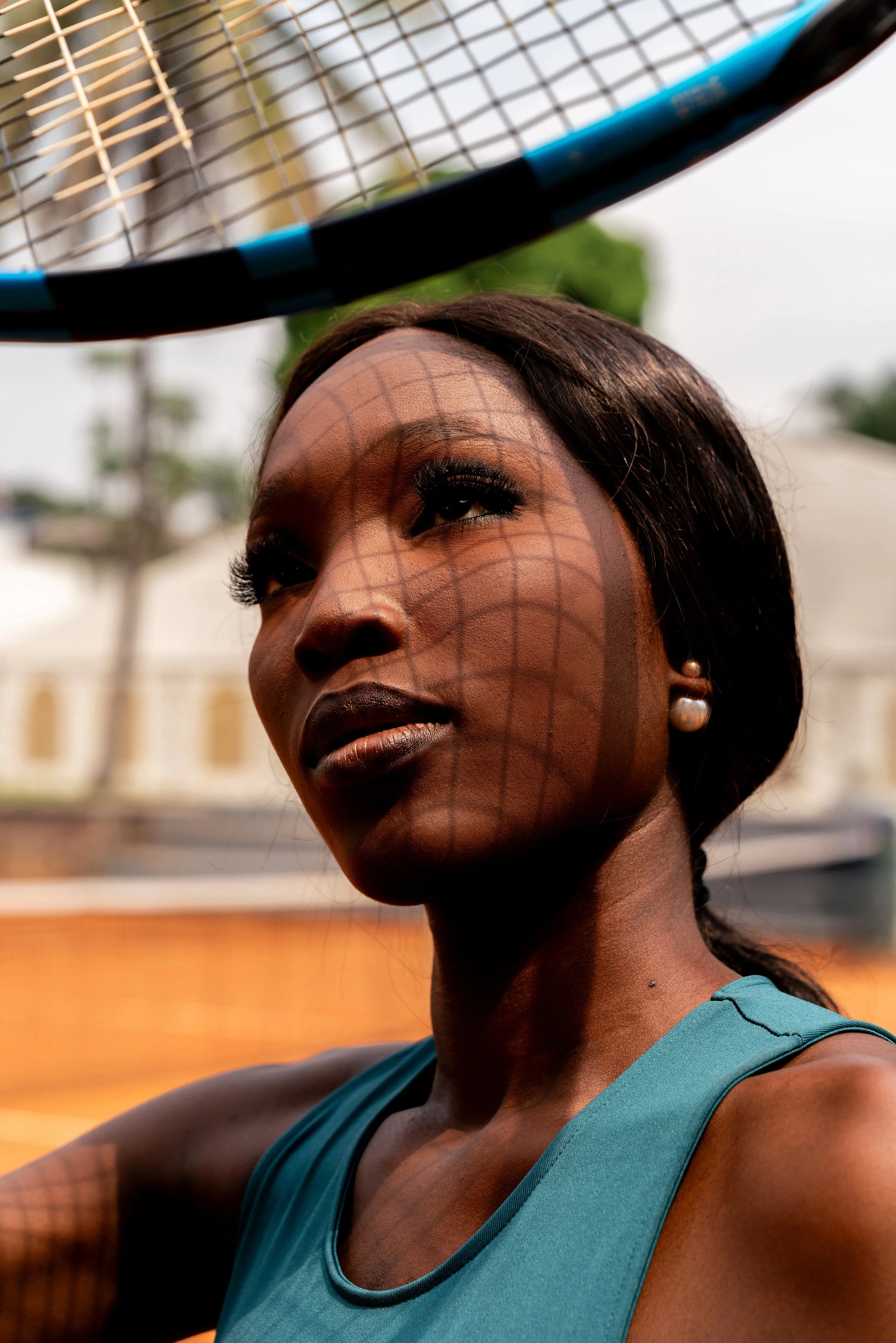 Une femme portant un débardeur bleu regarde vers le haut, le soleil crée une ombre en forme de filet sur son visage, avec un court court de tennis en arrière-plan.