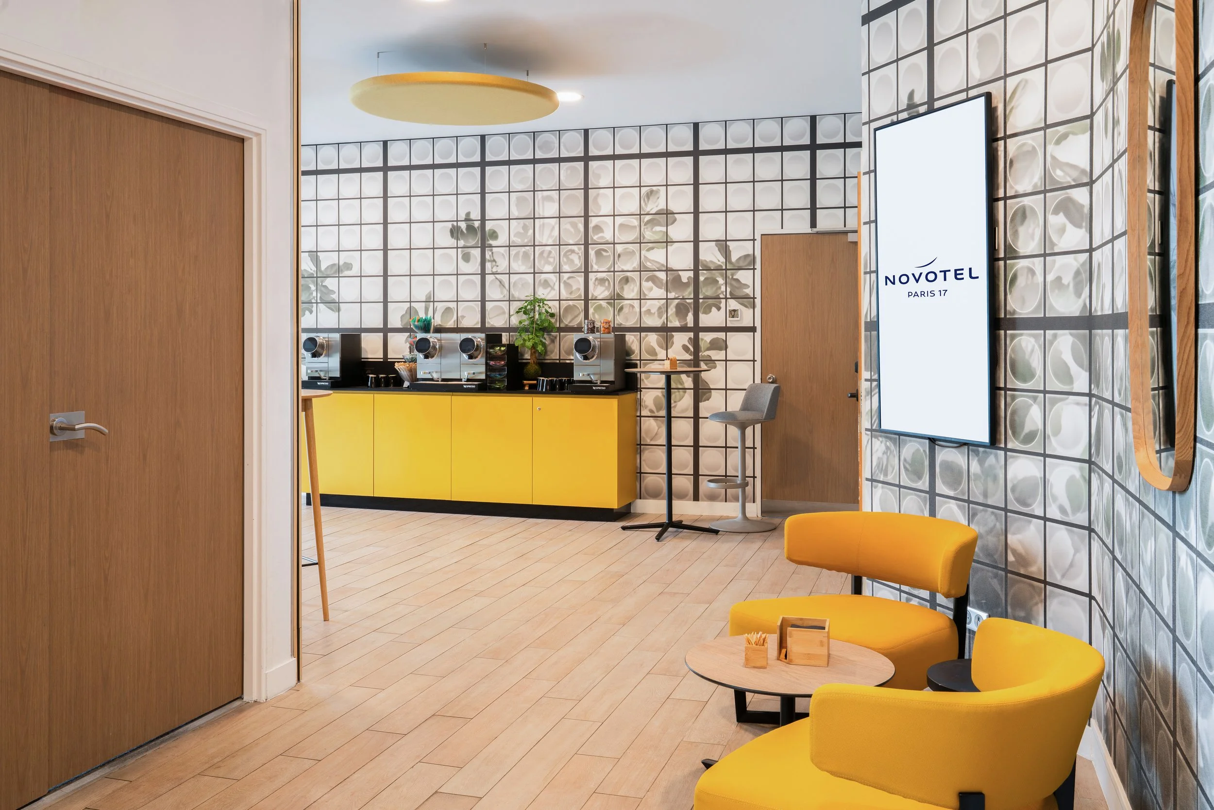 Salle de petit-déjeuner d'un hôtel Novotel avec murs décoratifs, fauteuils jaunes, un comptoir de café lumineux, et un écran numérique.