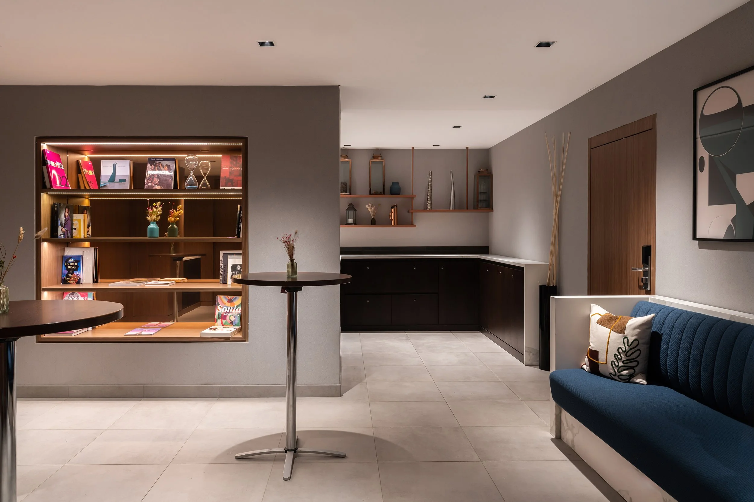 Salon moderne avec canapé bleu, étagère en bois avec livres et décorations, petit bar avec coussins à motifs, tableau abstrait sur le mur, et coin café en arrière-plan.