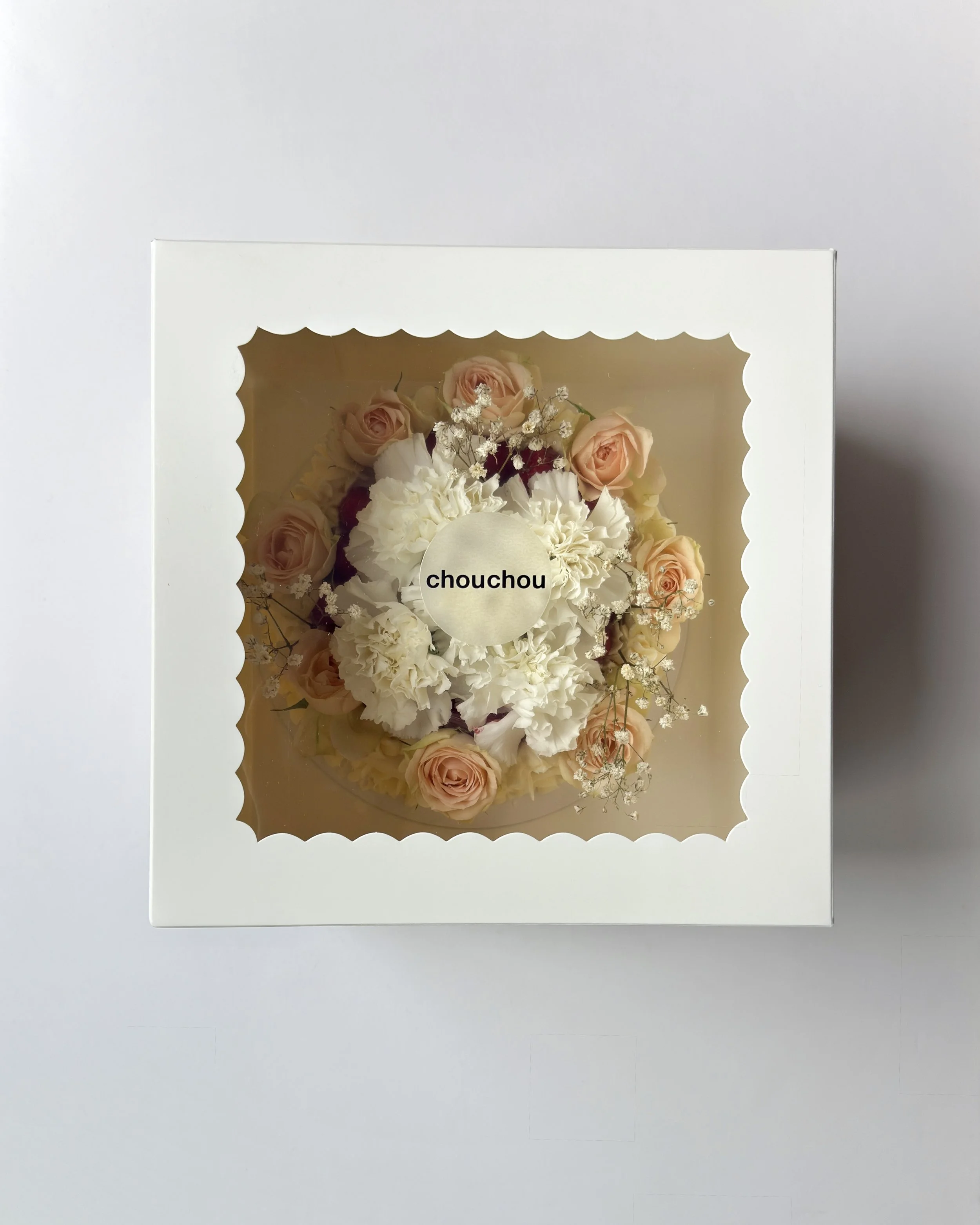 A boxed floral arrangement of white and light peach roses and white carnations, with a round label that says "chouchou" in the center, viewed from above.