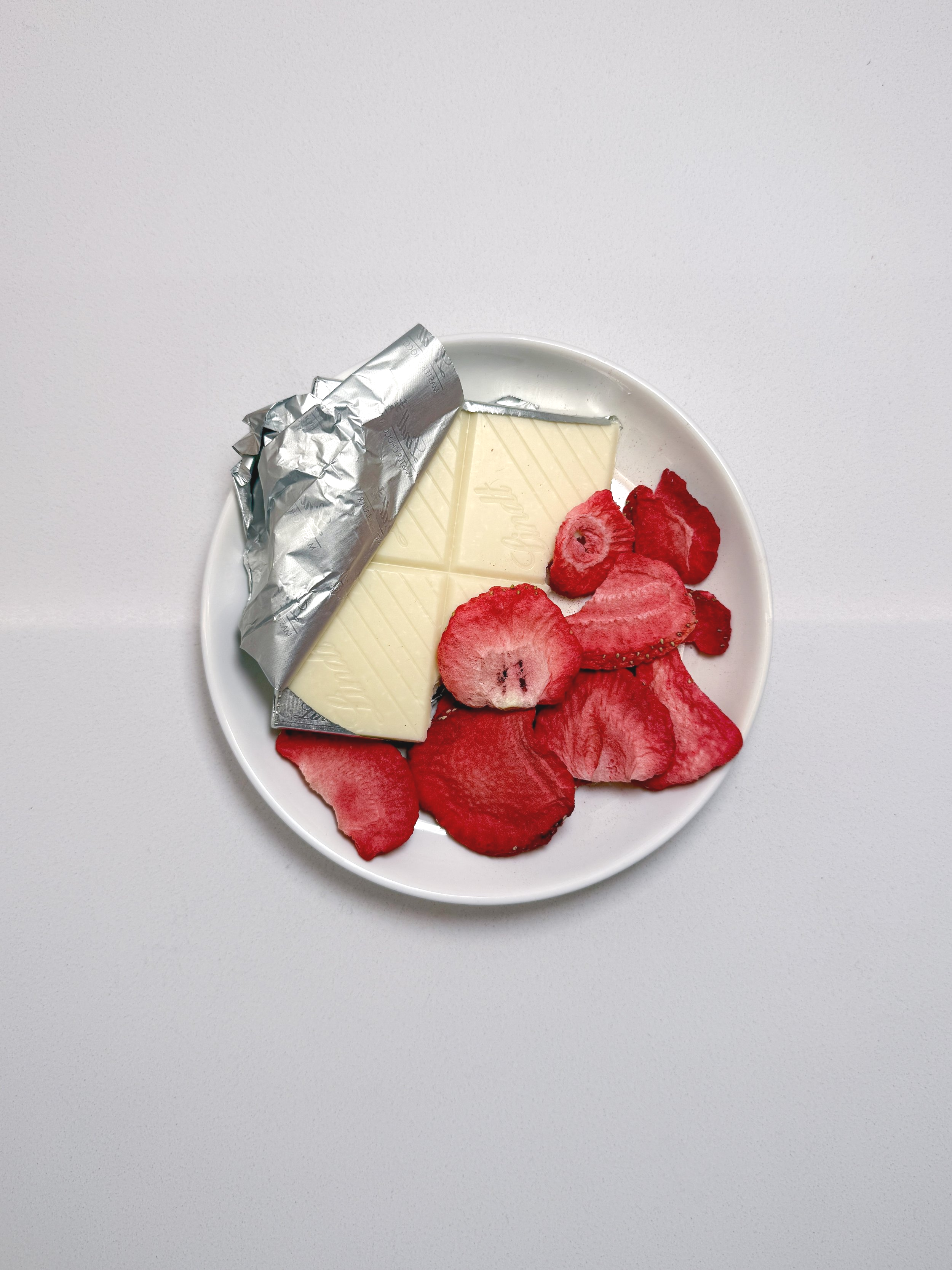 A white plate with a partially unwrapped white chocolate bar and red strawberry and raspberry slices.