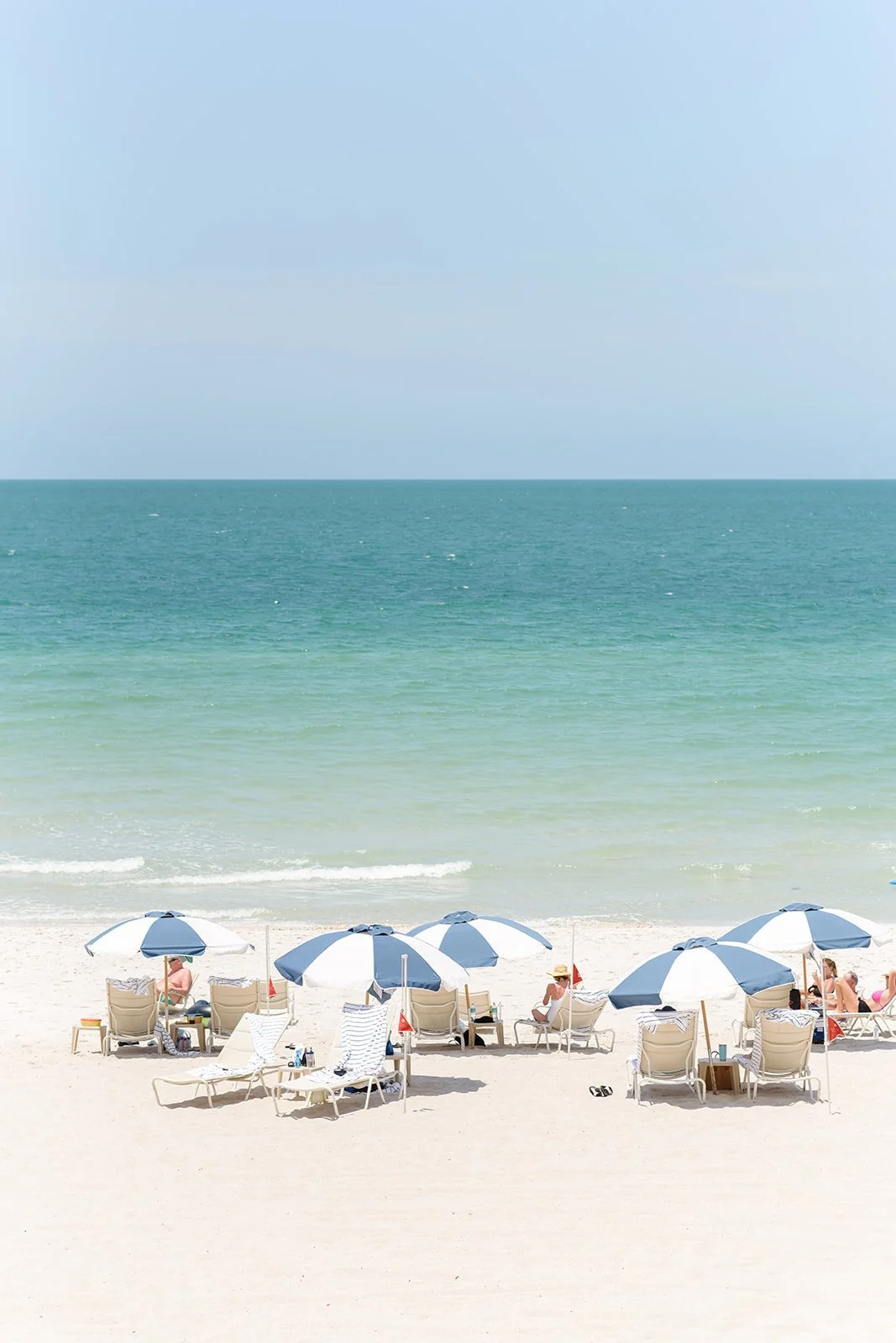 People relaxing on the beach under blue and white umbrellas, with the ocean and sky in the background.