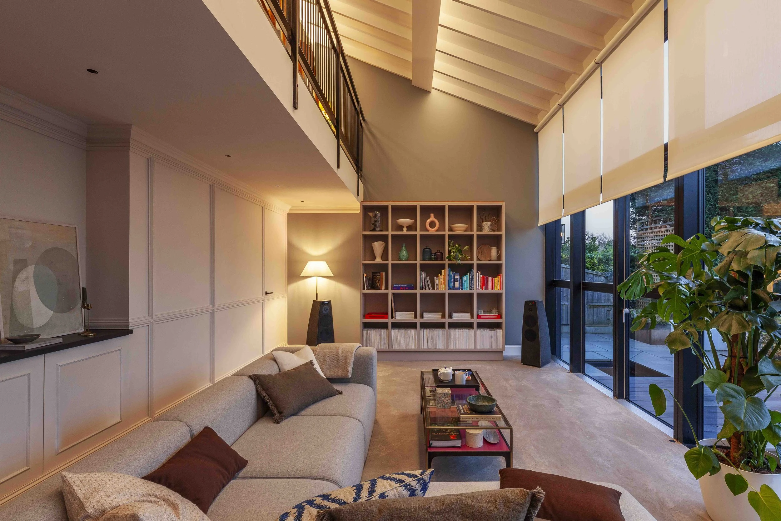 Modern living room with a large sectional sofa, a wooden bookshelf, large windows with roller shades, a coffee table, and a plant. A floor lamp provides warm lighting.