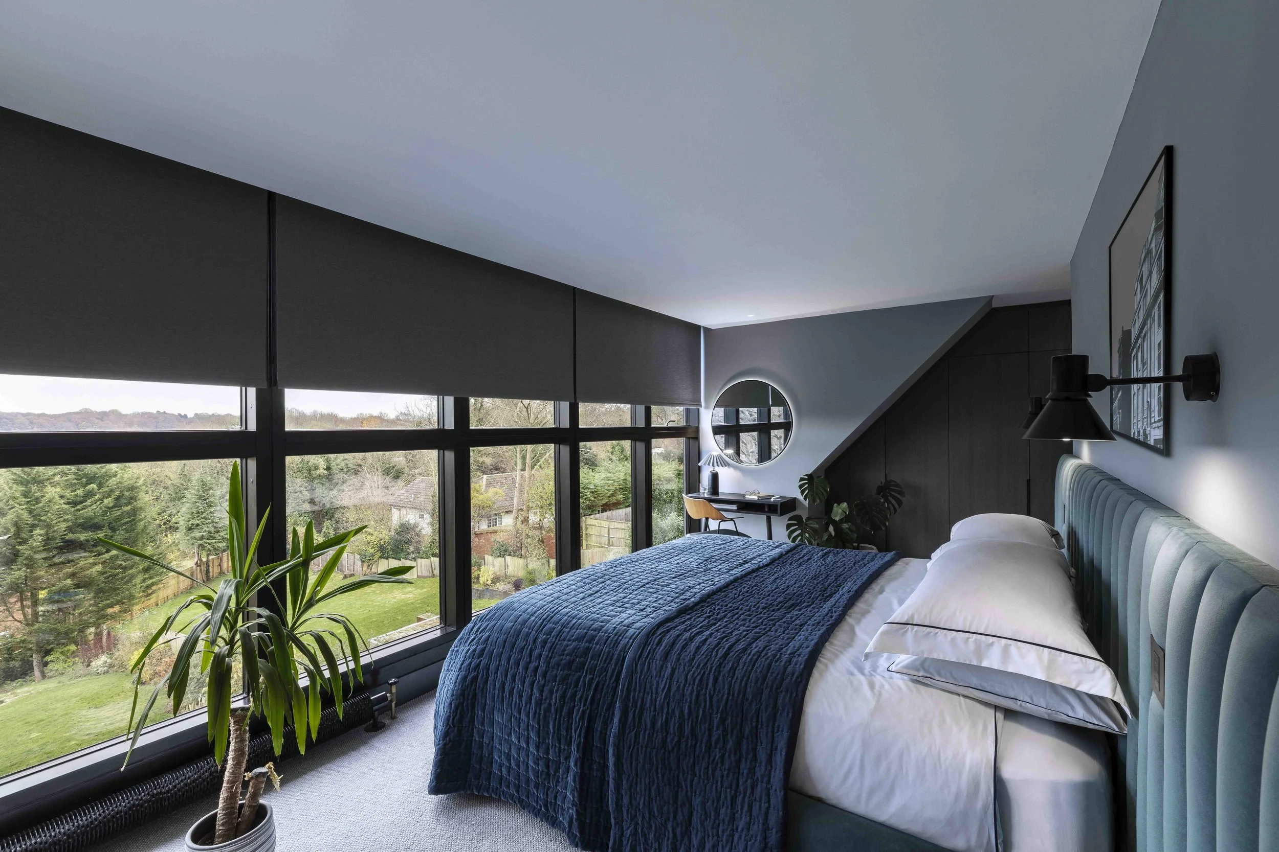 Modern bedroom with large windows showing outdoor landscape, a blue bed with white linens, a potted plant, and a small desk with a round mirror in the corner.