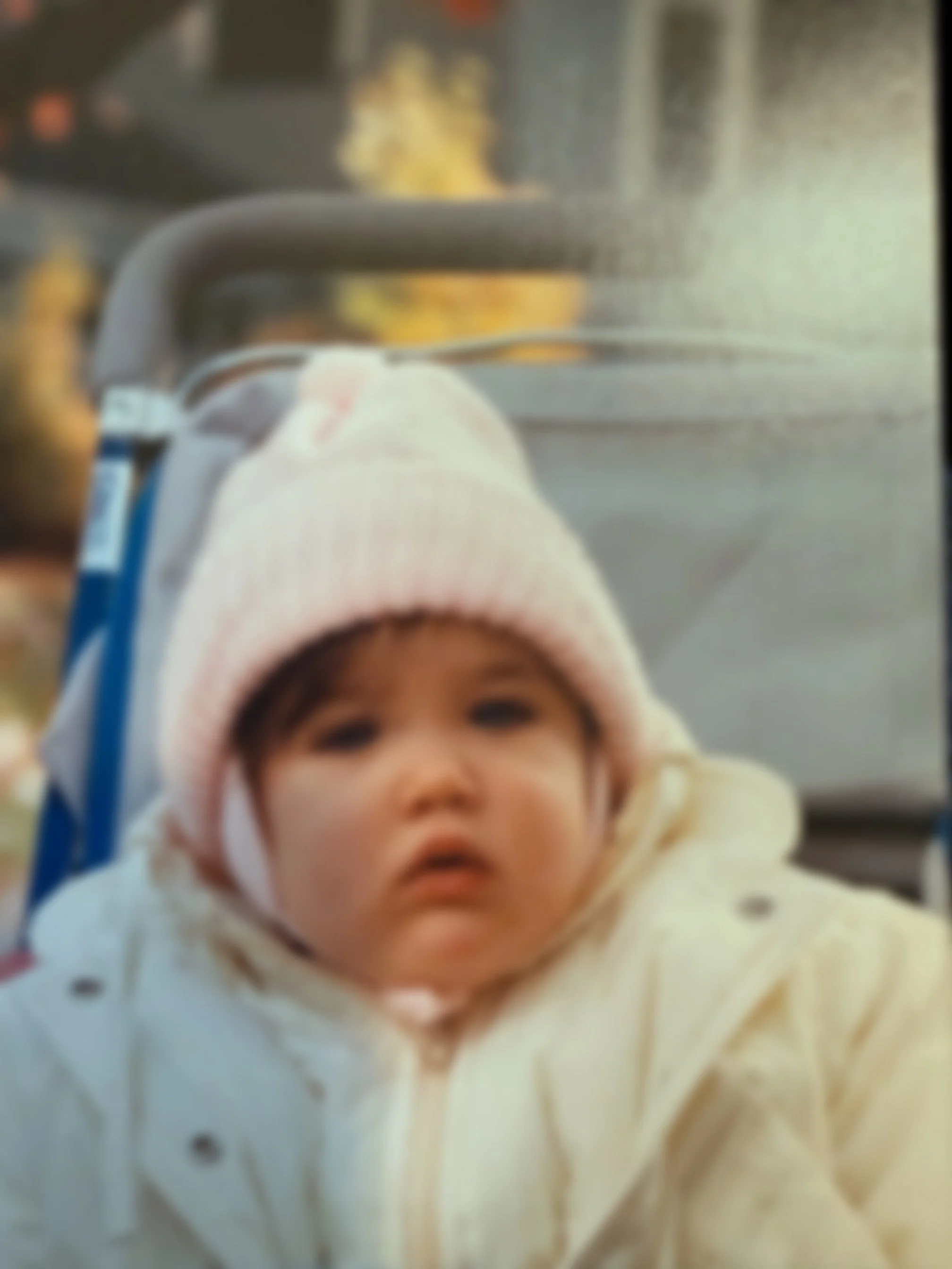 A young child wearing a pink knit hat and a cream-colored winter coat sitting in a stroller, looking directly at the camera, with a blurred outdoor background.