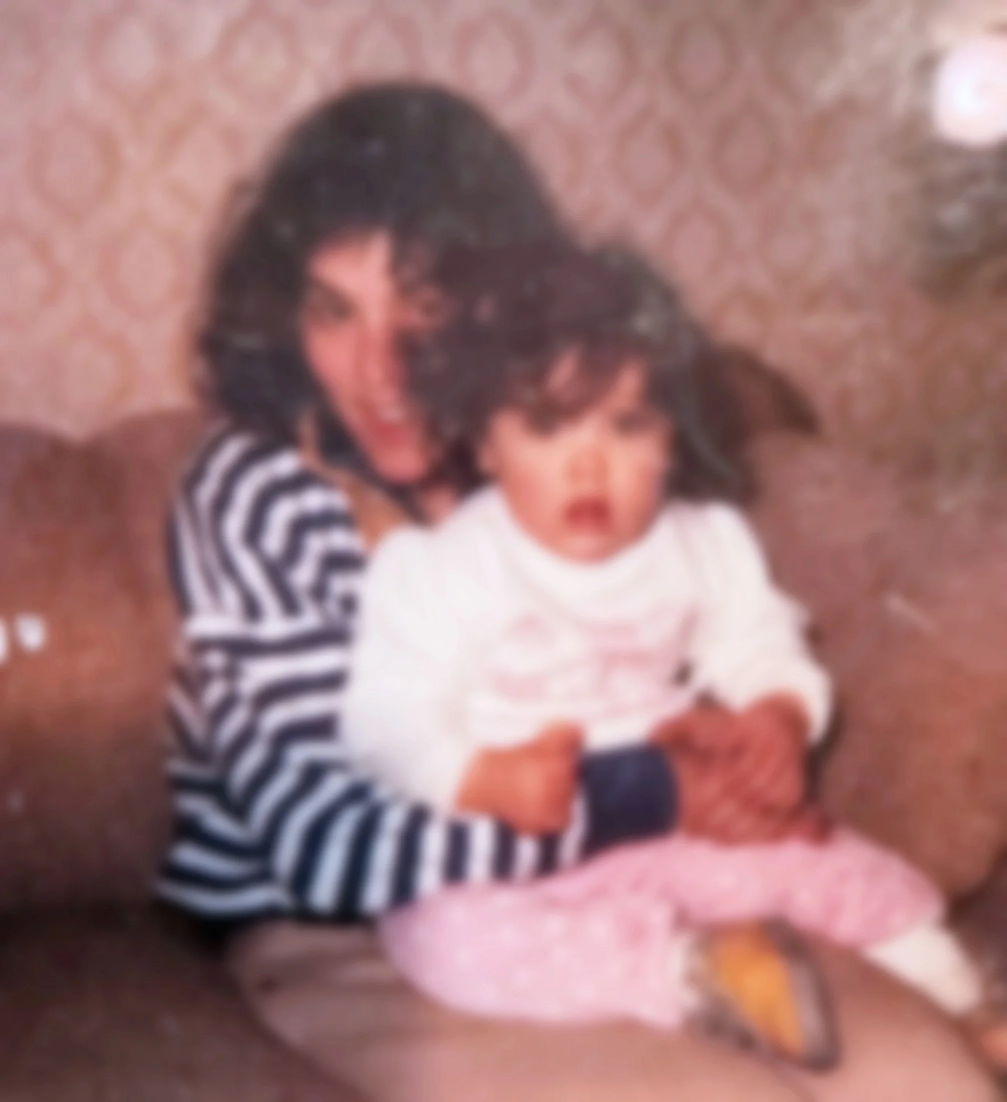 A woman with dark, curly hair and a striped shirt hugging a young girl with curly hair, wearing a white top and pink pants, sitting on a brown couch in a room with patterned wallpaper.