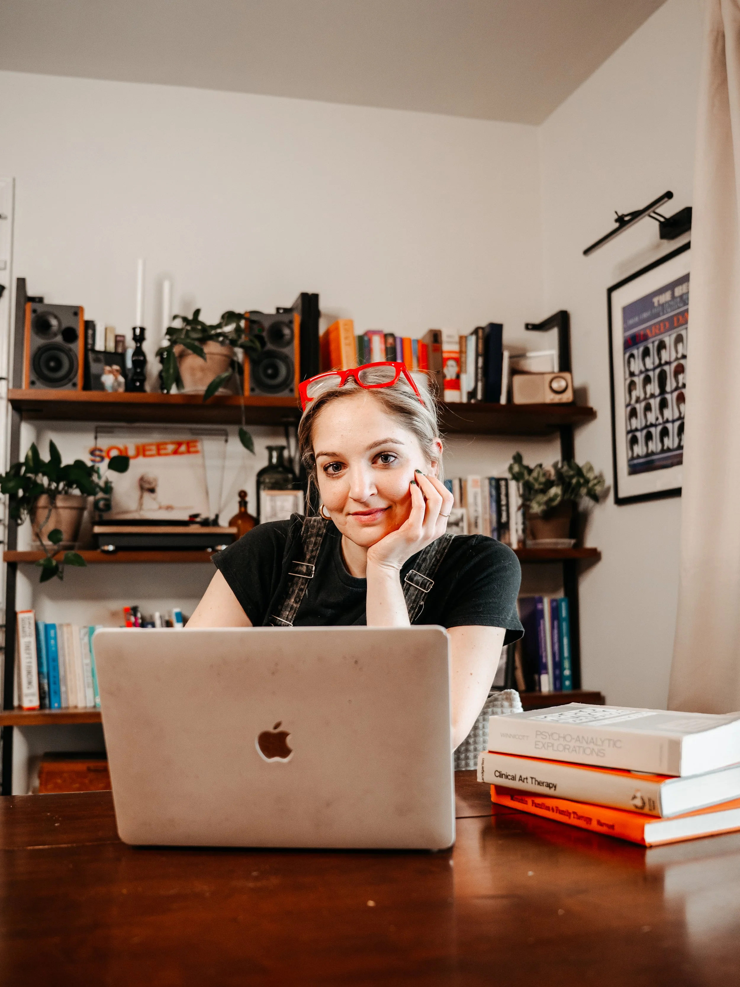 A woman with red glasses resting on her head, sitting at a wooden table with a silver MacBook, surrounded by books and plants in a cozy room.