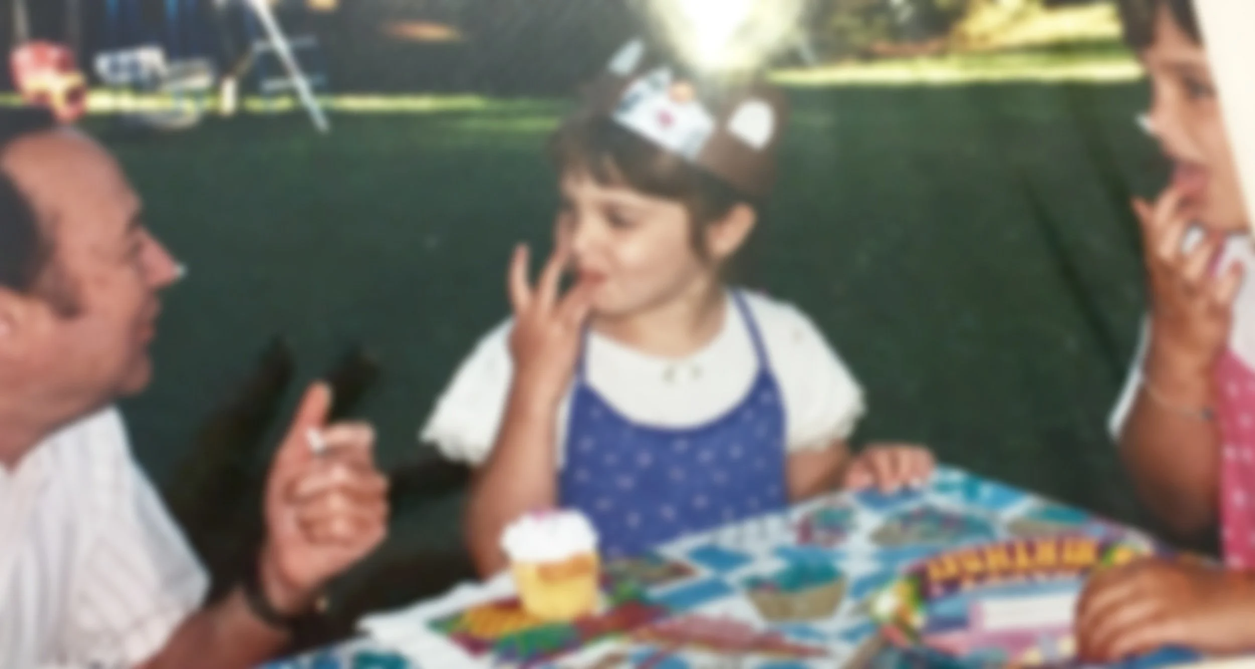 A young girl wearing a birthday crown and blue apron is at a birthday party outside, touching her face near her lips. Two adults are sitting next to her, one man on the left holding a phone and the woman on the right eating. The table is covered with colorful party tablecloth, cupcakes, and party favors. In the background, there is a grassy area and some outdoor play equipment.