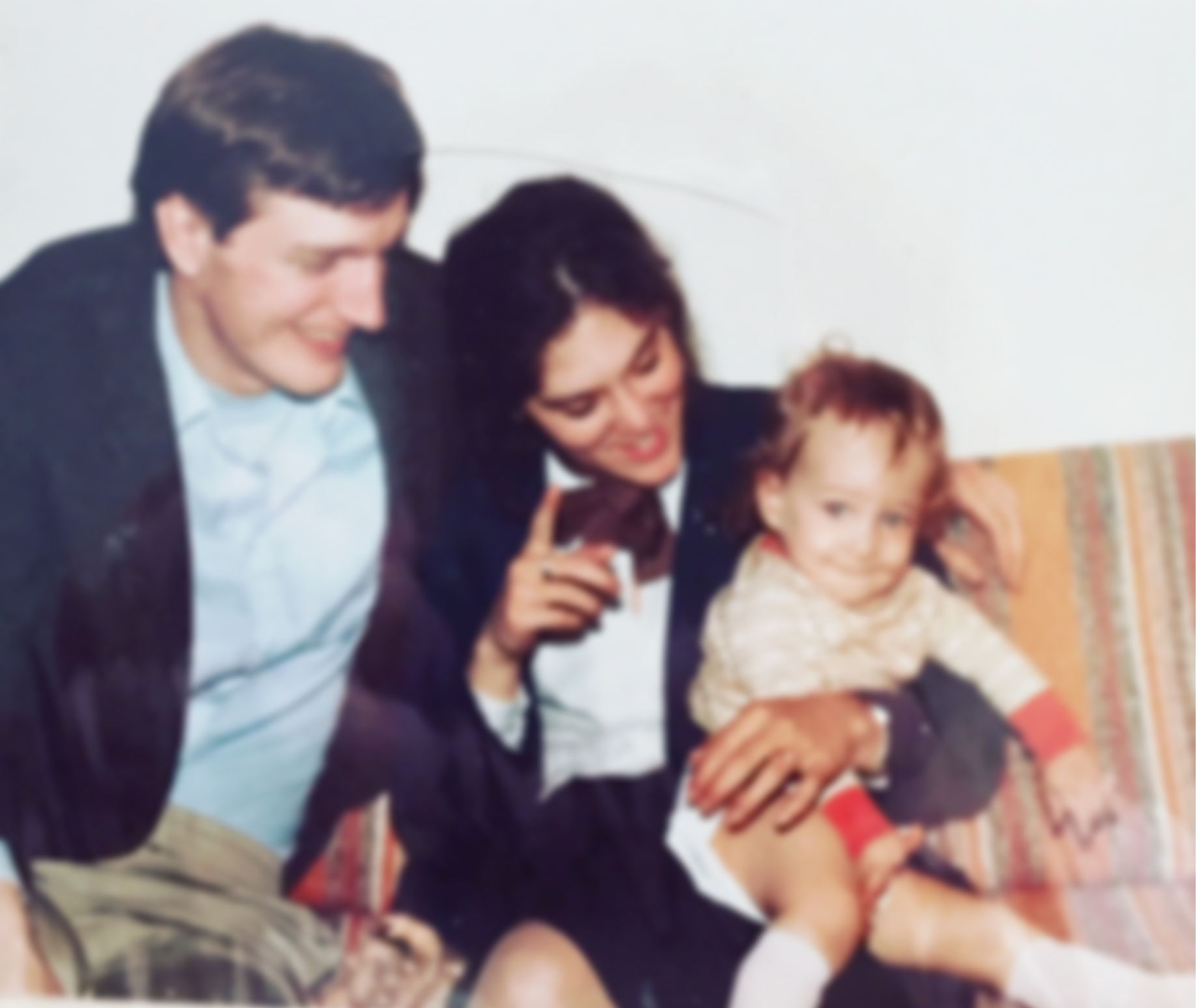 Three people sitting on a patterned couch, smiling and posing, including two adults and a young boy with a tan coat and shorts.