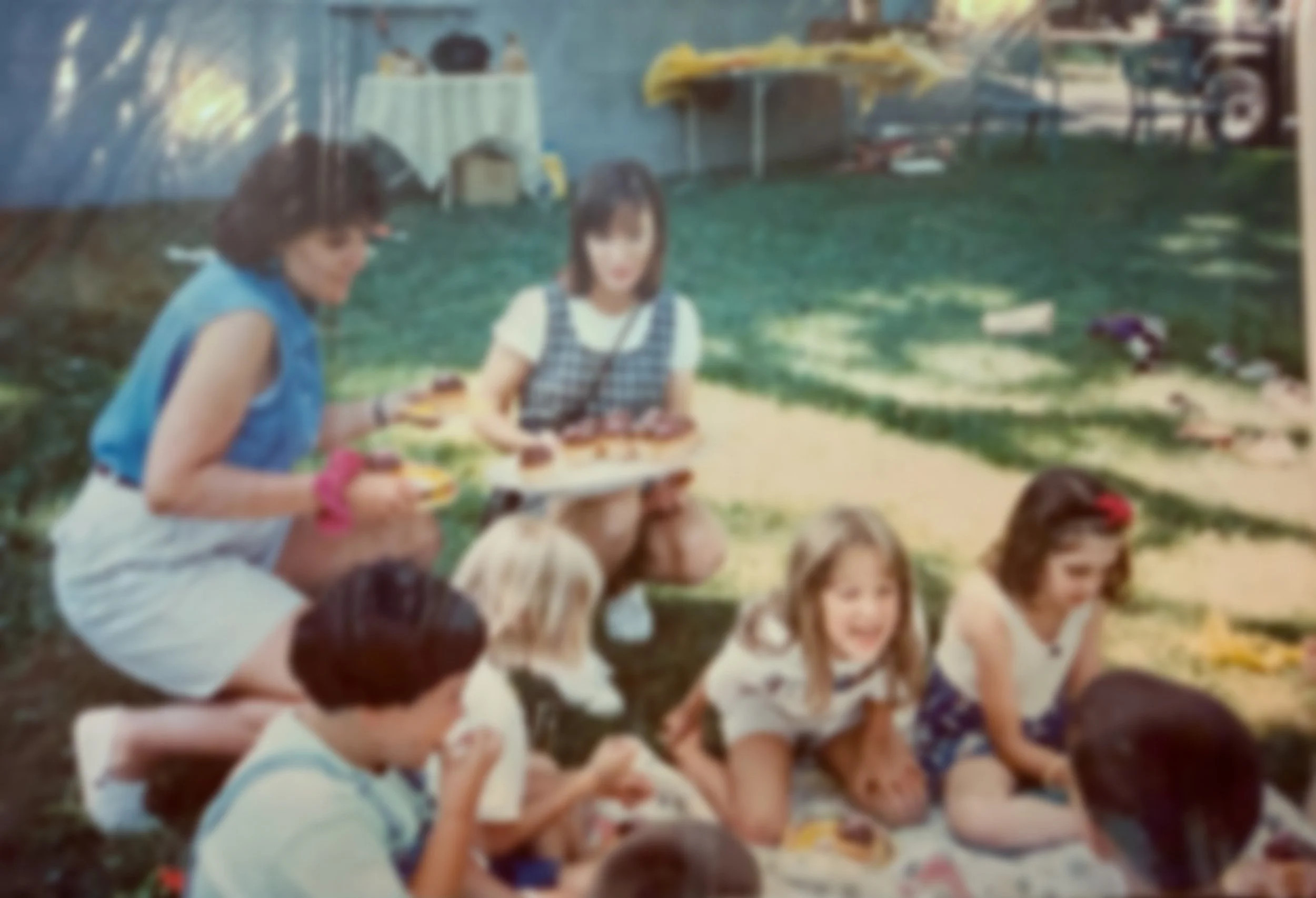 Children and adults having a picnic outdoors, with some eating and one person holding a tray of grilled food on a sunny day.