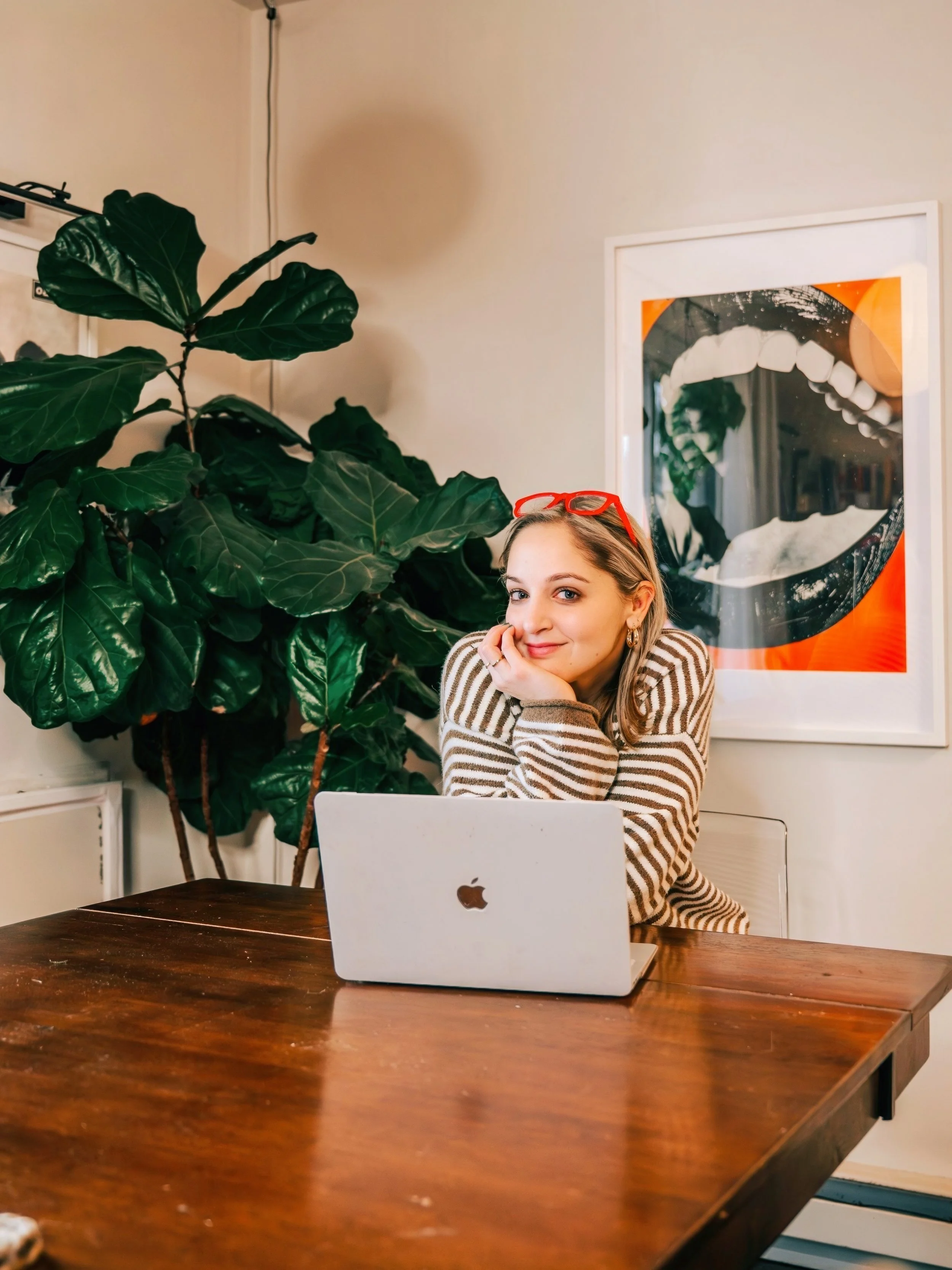 A young woman with blonde hair and red glasses resting her chin on her hand, sitting at a wooden table with an open silver MacBook in front of her. She is smiling and wearing a striped sweater. Behind her is a large green leafy plant and a colorful framed print of an open mouth with teeth.