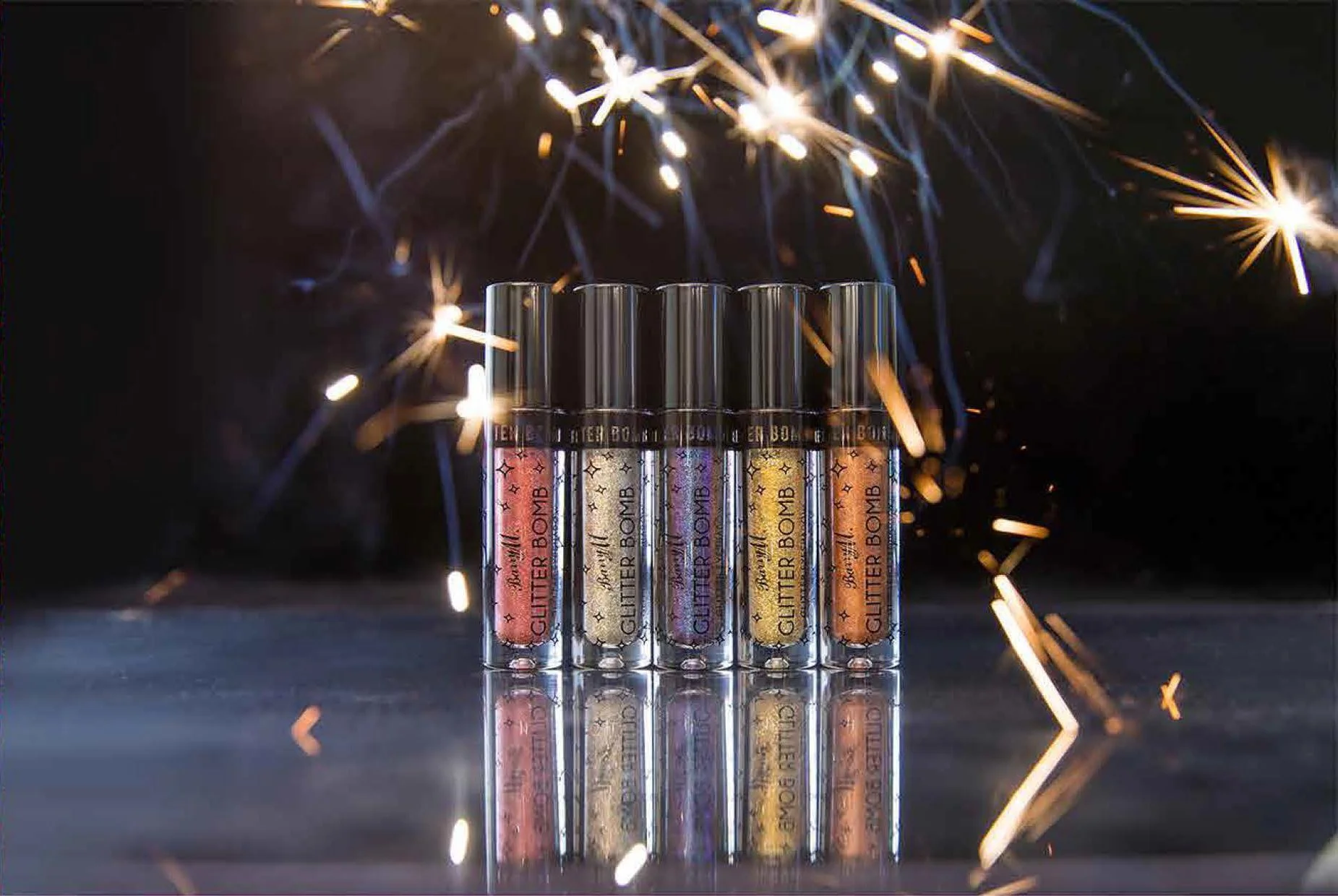 Five tubes of glitter lip gloss arranged in a row with sparklers in the background illuminating the scene.