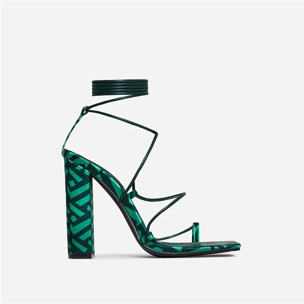 A high-heeled sandal with a patterned block heel, green geometric design, and thin black straps, set against a light background.