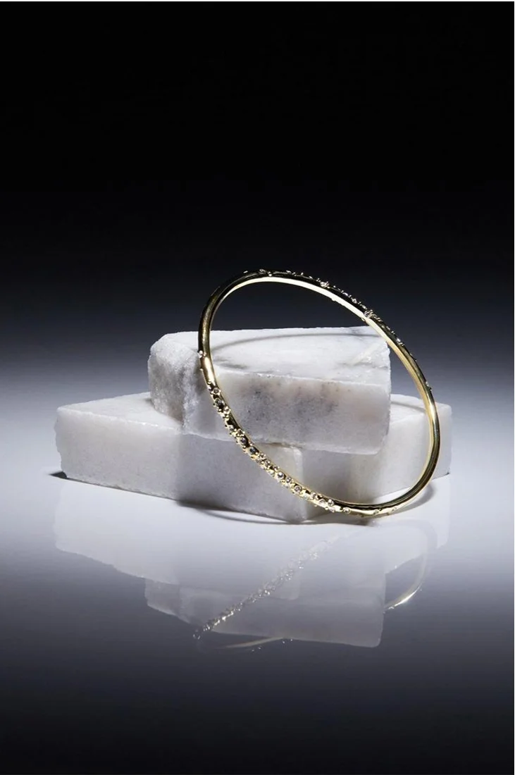 A gold bracelet with small gemstones resting on three white marble blocks against a dark background.