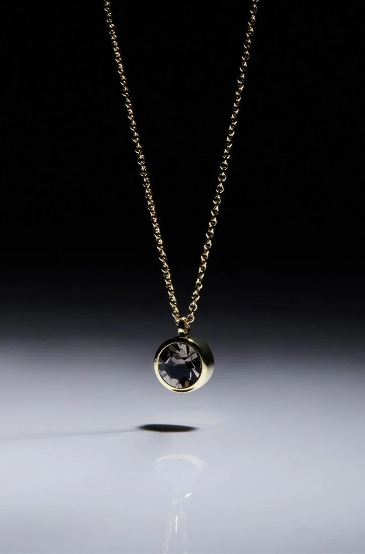 Gold necklace with a single round black gemstone pendant hanging in mid-air with soft shadow below.