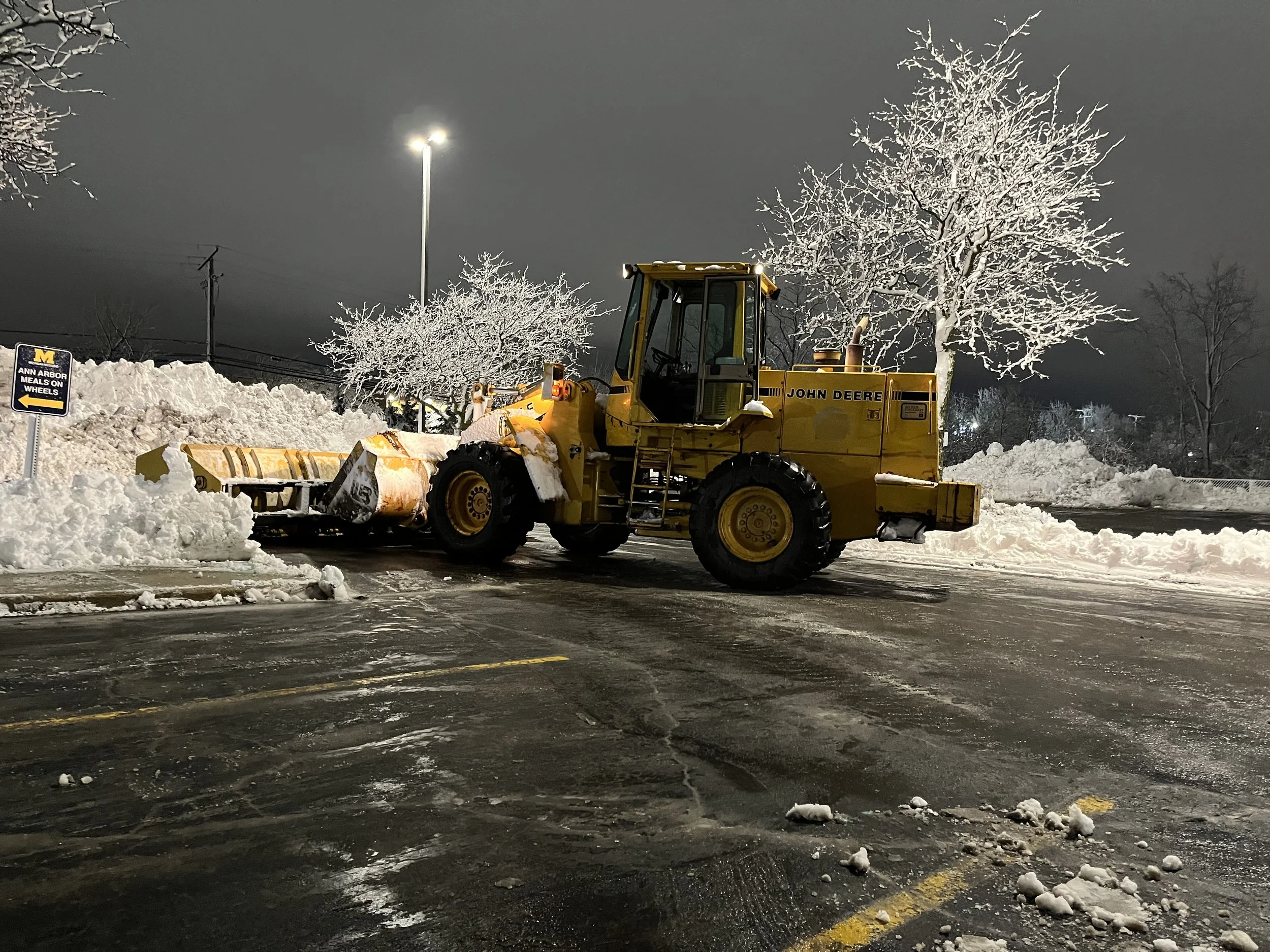 A yellow John Deere snowplow clearing snow from a parking lot during winter at night. Snow piles and trees covered in snow are visible in the background, along with a lamppost and a sign.