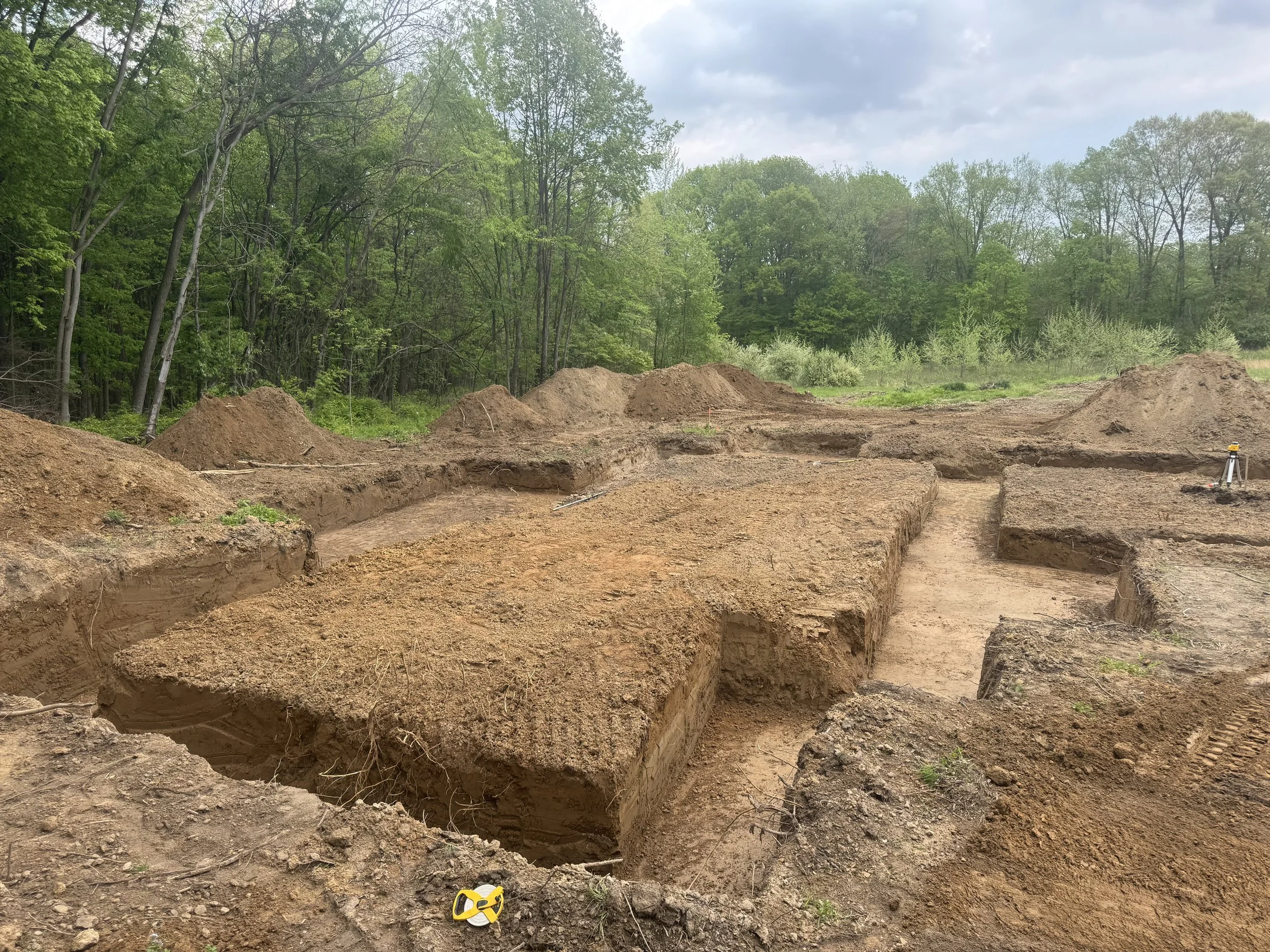 Excavated construction site with trenches and mounds of dirt, surrounded by green trees and under a partly cloudy sky.