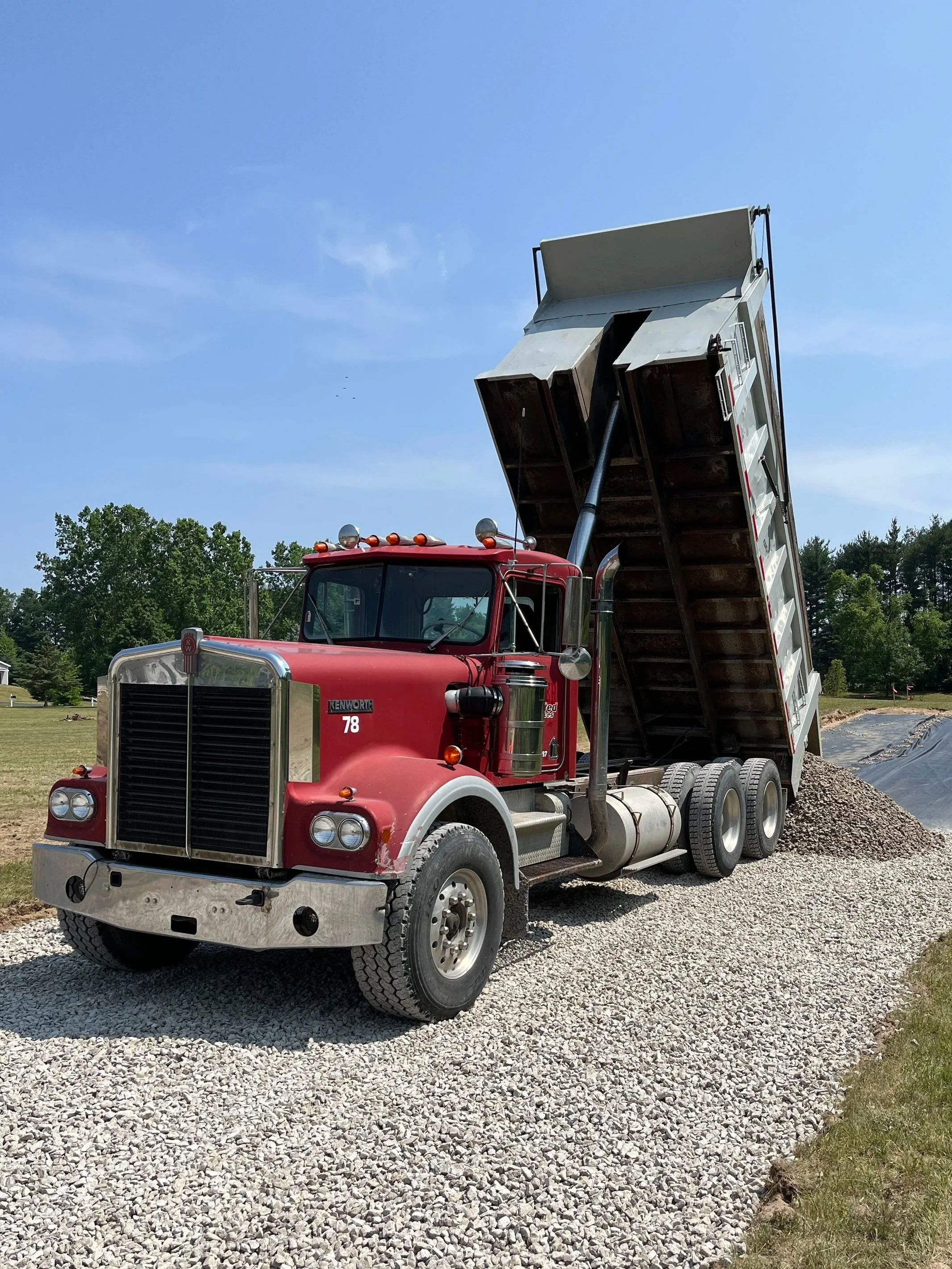 Red dump truck with raised bed on gravel surface, blue sky, green trees in background.