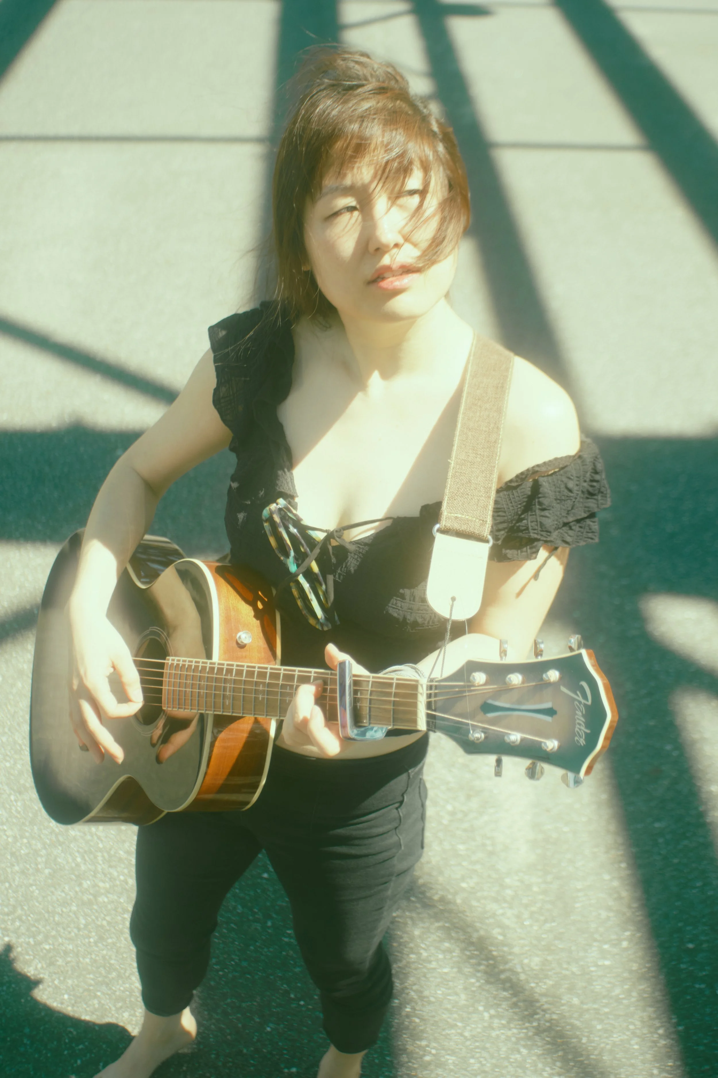 A woman with shoulder-length hair playing an acoustic guitar outdoors on a sunny day, wearing a black off-shoulder top and black pants. JSnaps Photography New York City Fashion lifestyle photography 