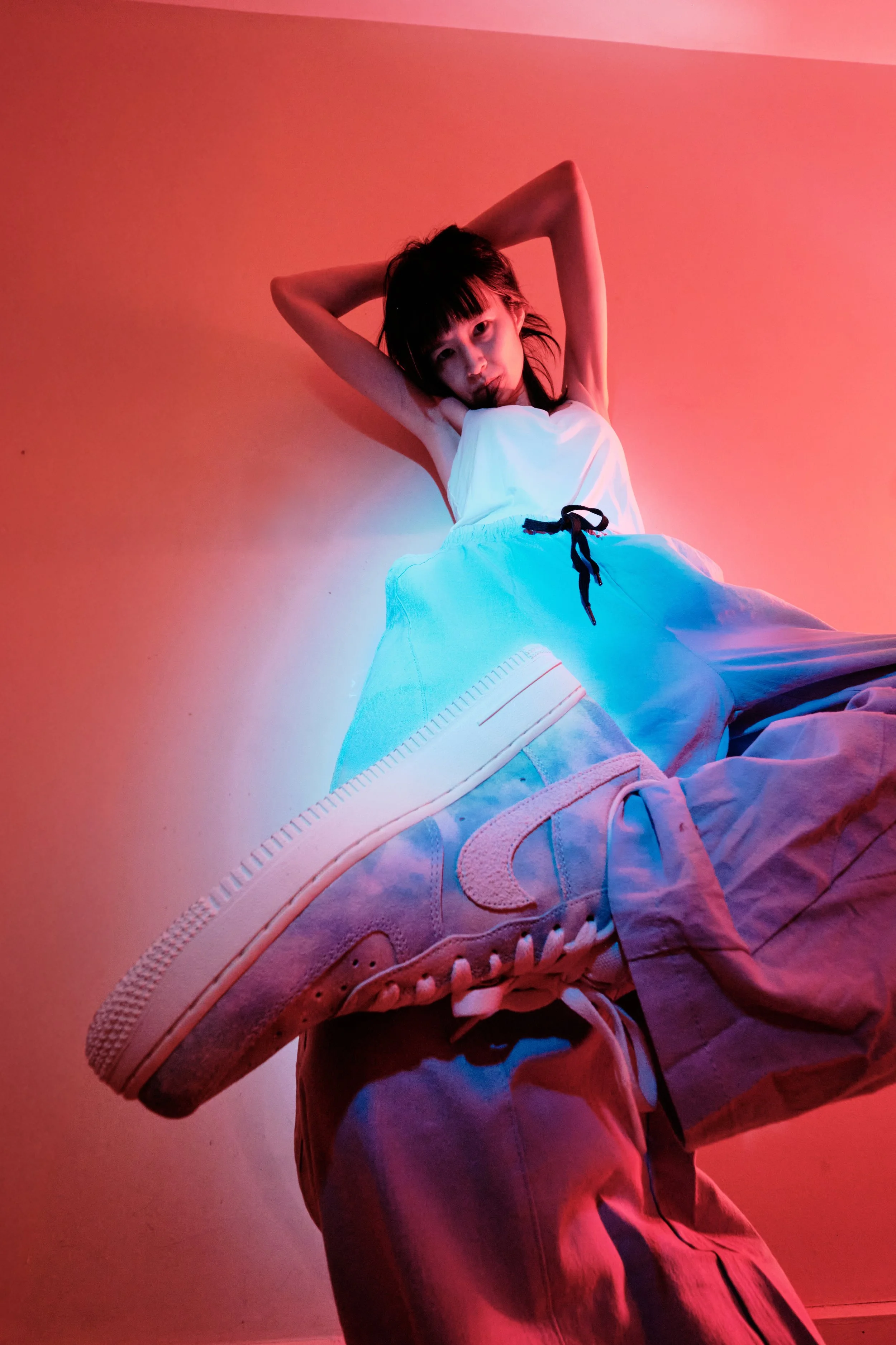A young woman with short dark hair leaning against a pink wall, wearing baggy pants and a white top, with one arm raised behind her head. She is posing with one foot close to the camera, showcasing a light-colored sneaker. The lighting is colorful, w