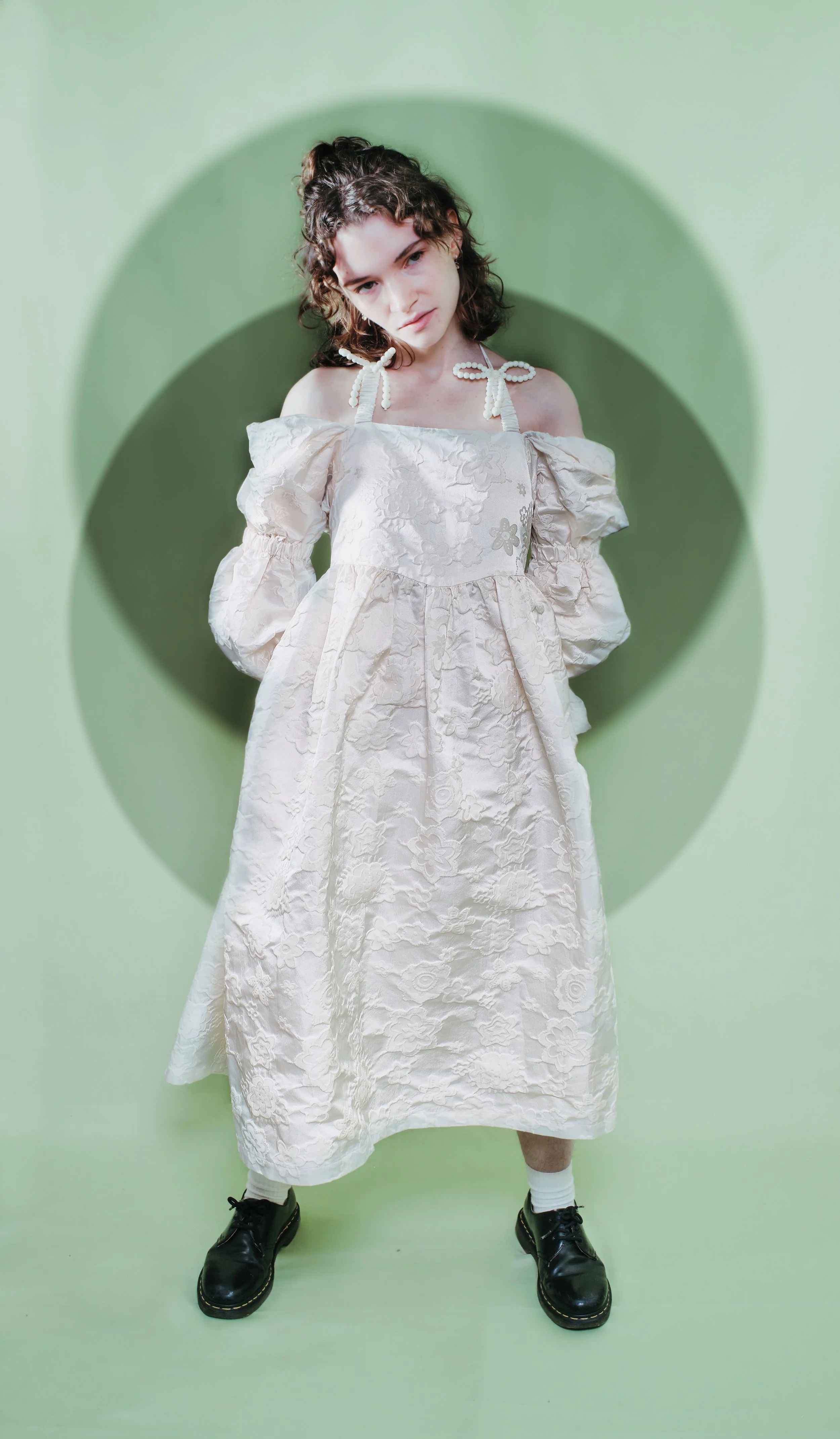 A young woman with curly hair wearing a floral lace dress with puffed sleeves, standing in front of a green background with circular shadows, wearing black shoes and white socks. Headshot of a female JSnaps Photography New York City Fashion lifestyle