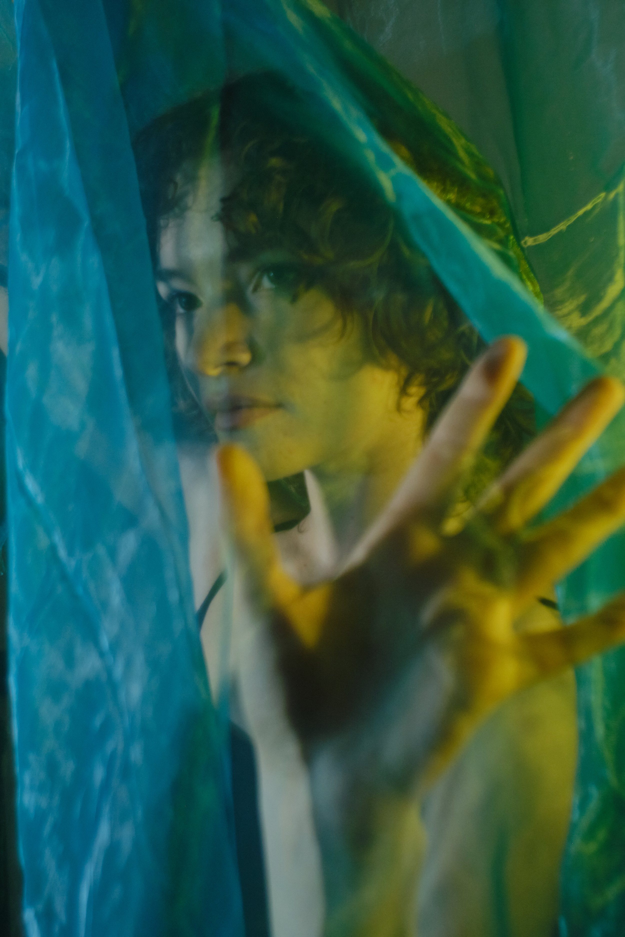 A woman with curly hair behind a translucent blue curtain, touching it with her hand as she looks through. Portrait Photographer New York City