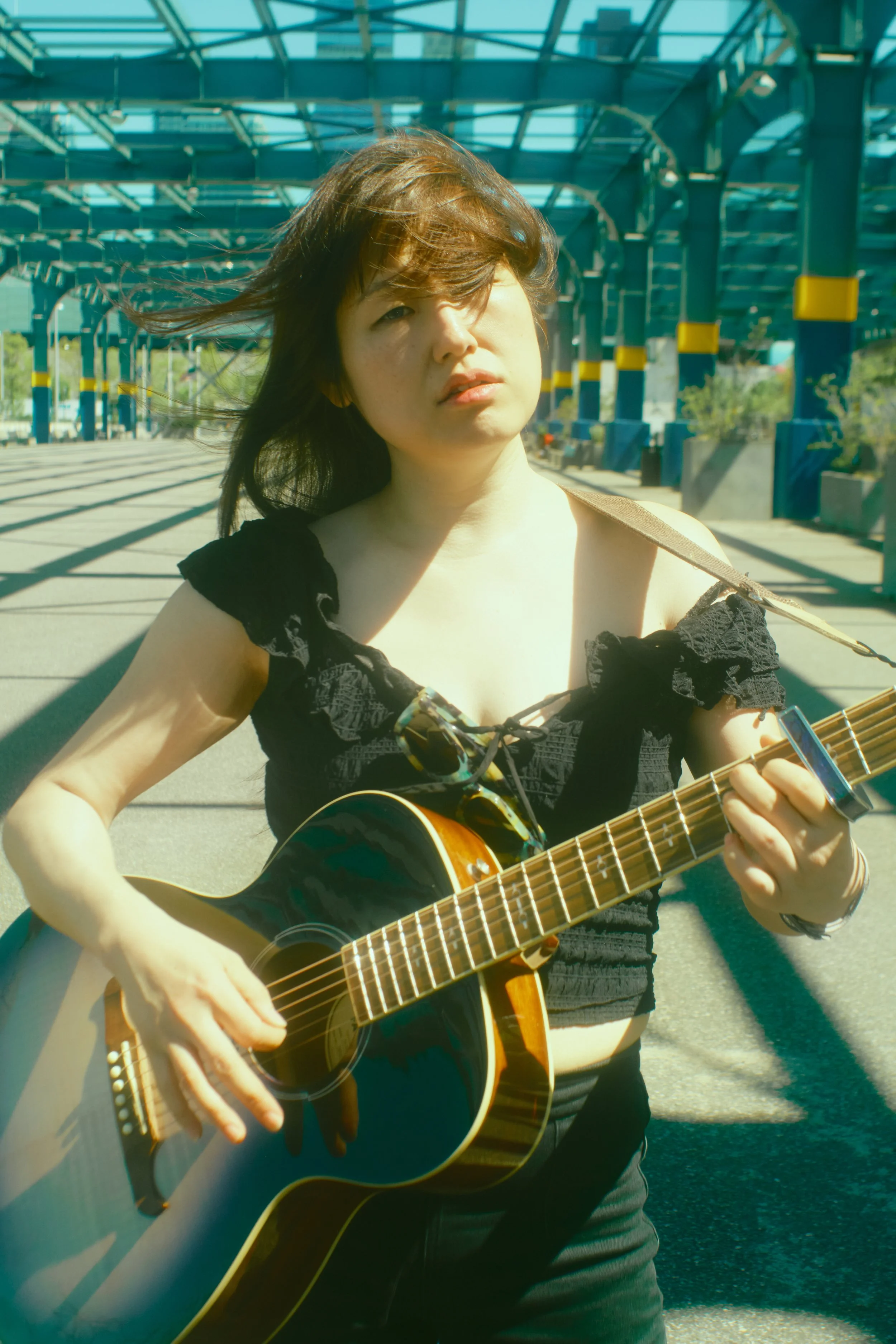 A woman with brown hair playing an acoustic guitar outdoors on a sunny day, standing under a large glass roof structure. JSnaps Photography New York City Fashion lifestyle photography 