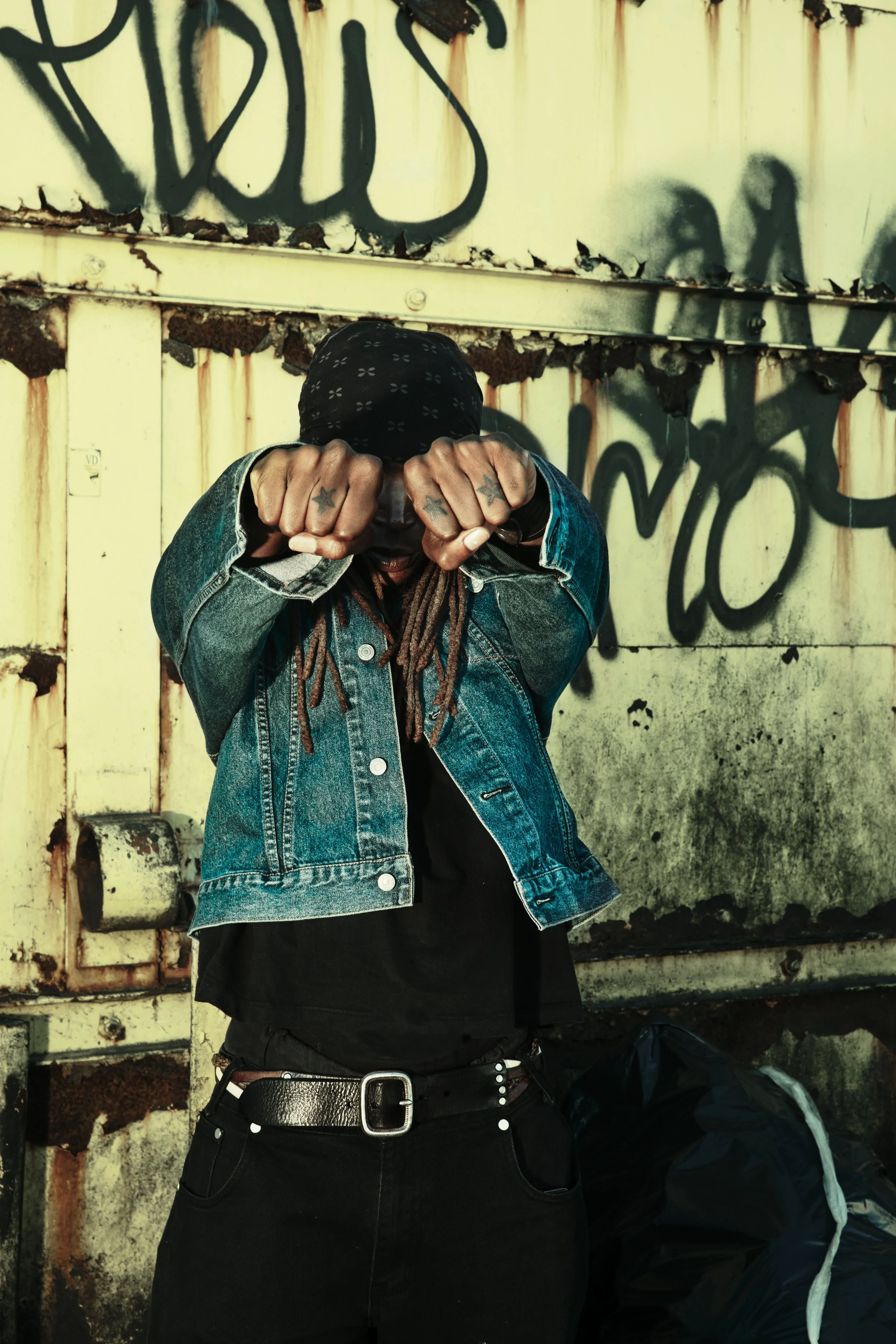 A man with dreadlocks wearing a bandana, denim jacket, and black t-shirt standing against a graffiti-covered, weathered wall in an urban setting. JSnaps Photography New York City Fashion lifestyle photography 