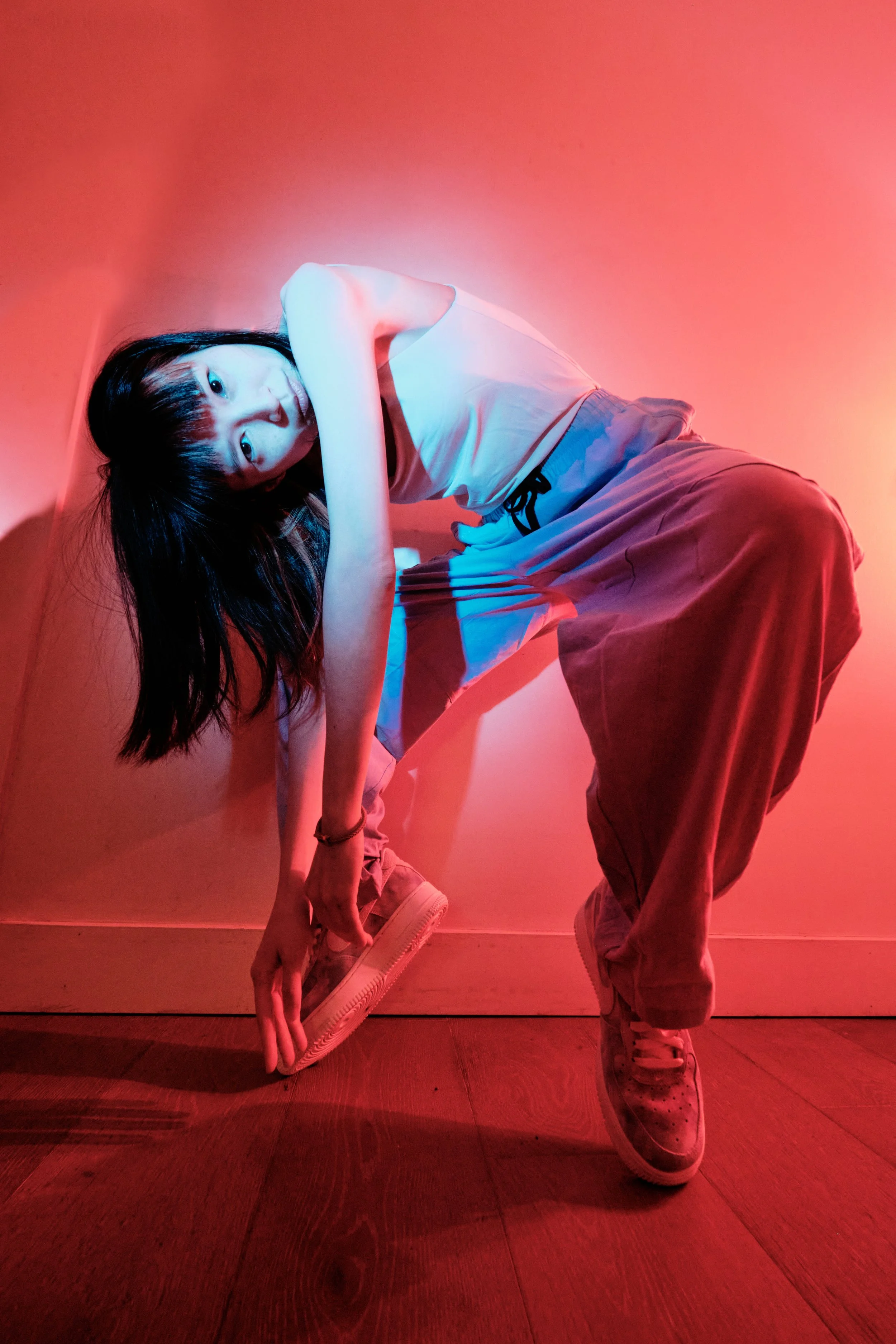 A young woman with long dark hair is crouching against a wall with red and blue lighting, holding her shoe while wearing casual white top, beige pants, and sneakers. JSnaps Photography New York City Fashion lifestyle photography 