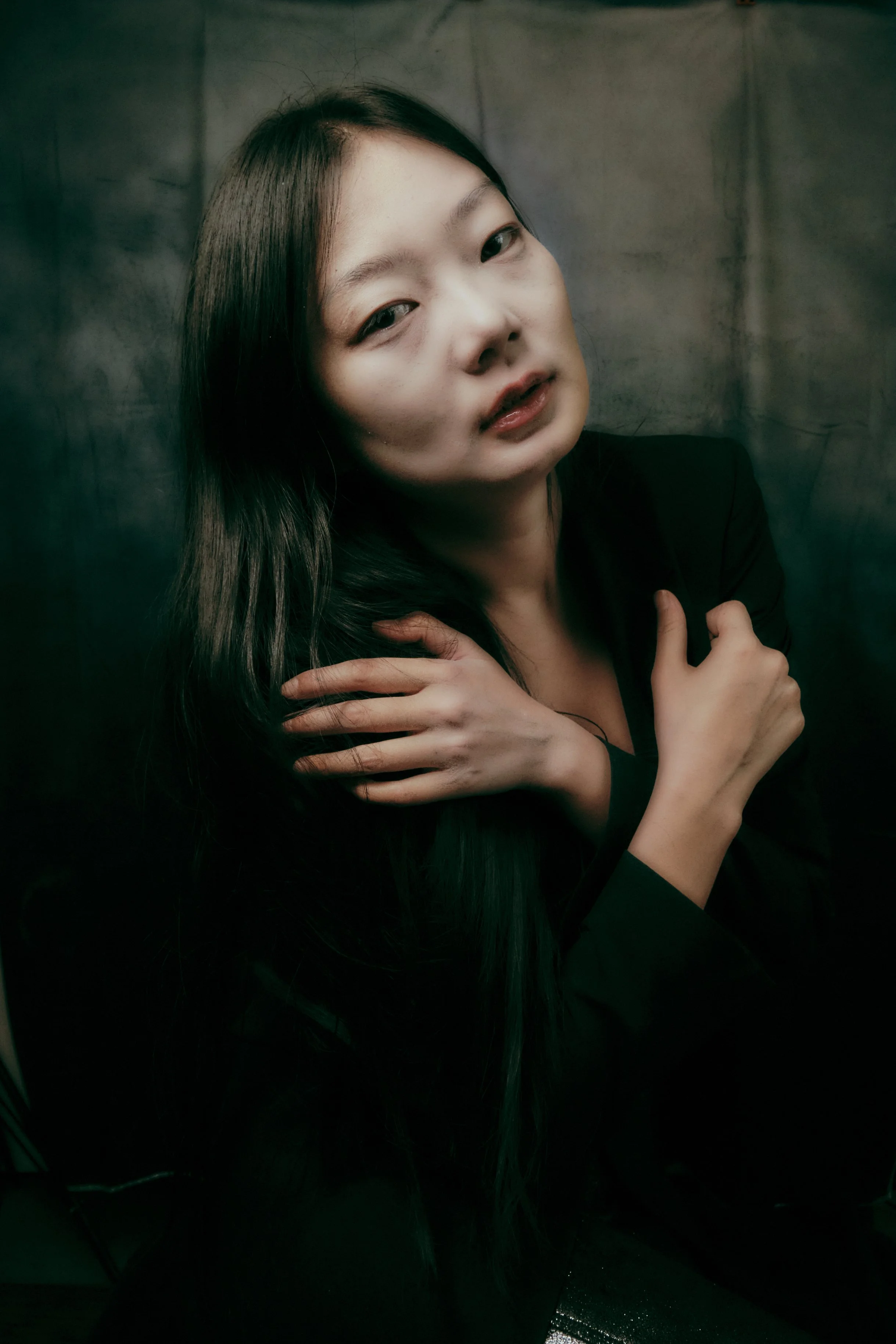A woman with long dark hair, wearing a black blazer, holding her arm across her chest, with a neutral expression, standing against a dark, textured background. Portrait Photographer New York City
