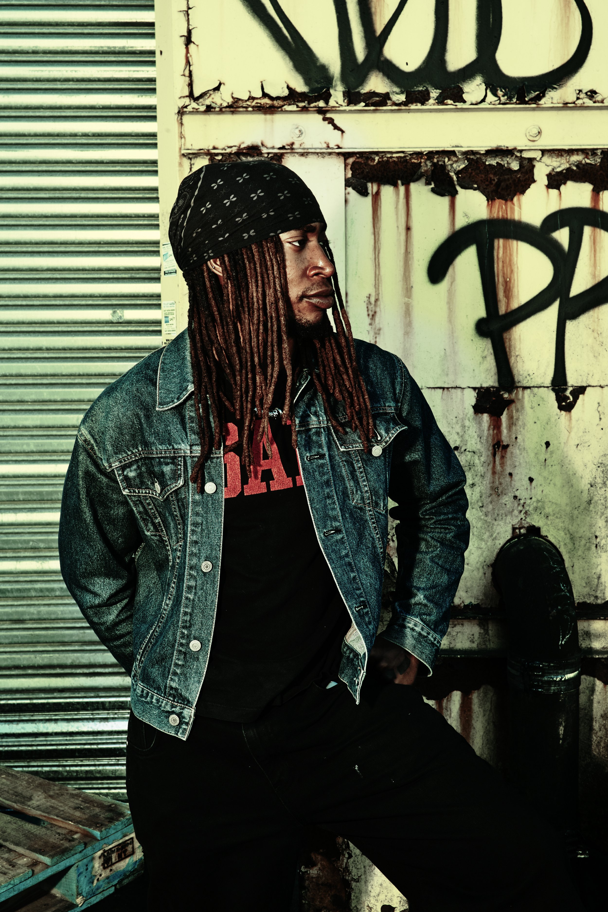 A man with dreadlocks wearing a bandana, denim jacket, and black t-shirt standing against a graffiti-covered, weathered wall in an urban setting. JSnaps Photography New York City Fashion lifestyle photography 