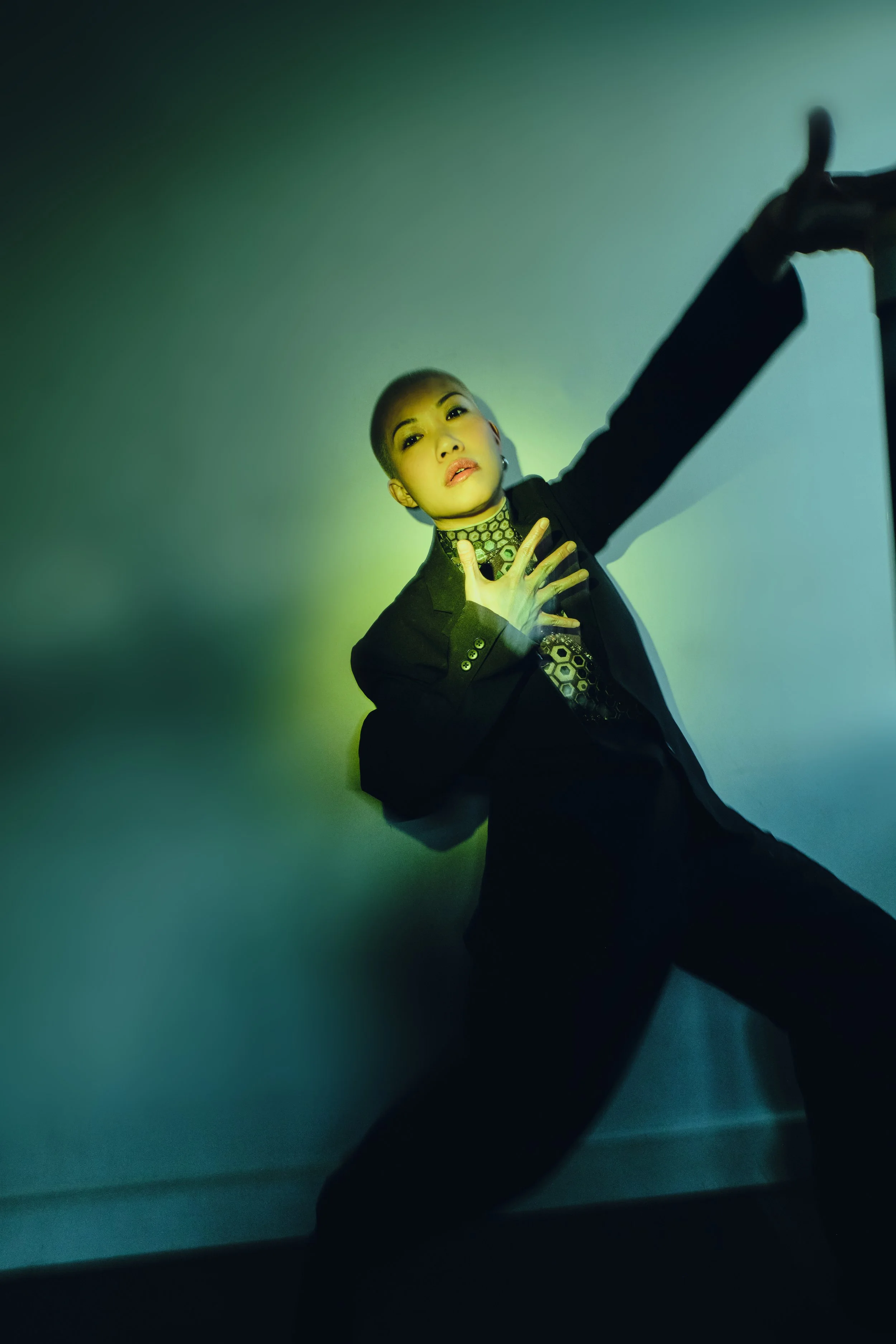 A person with a shaved head posing against a gradient background, wearing a black suit with a patterned shirt, and making a dramatic gesture with one hand on their chest and the other extended upward. JSnaps Photography New York City Fashion lifestyl