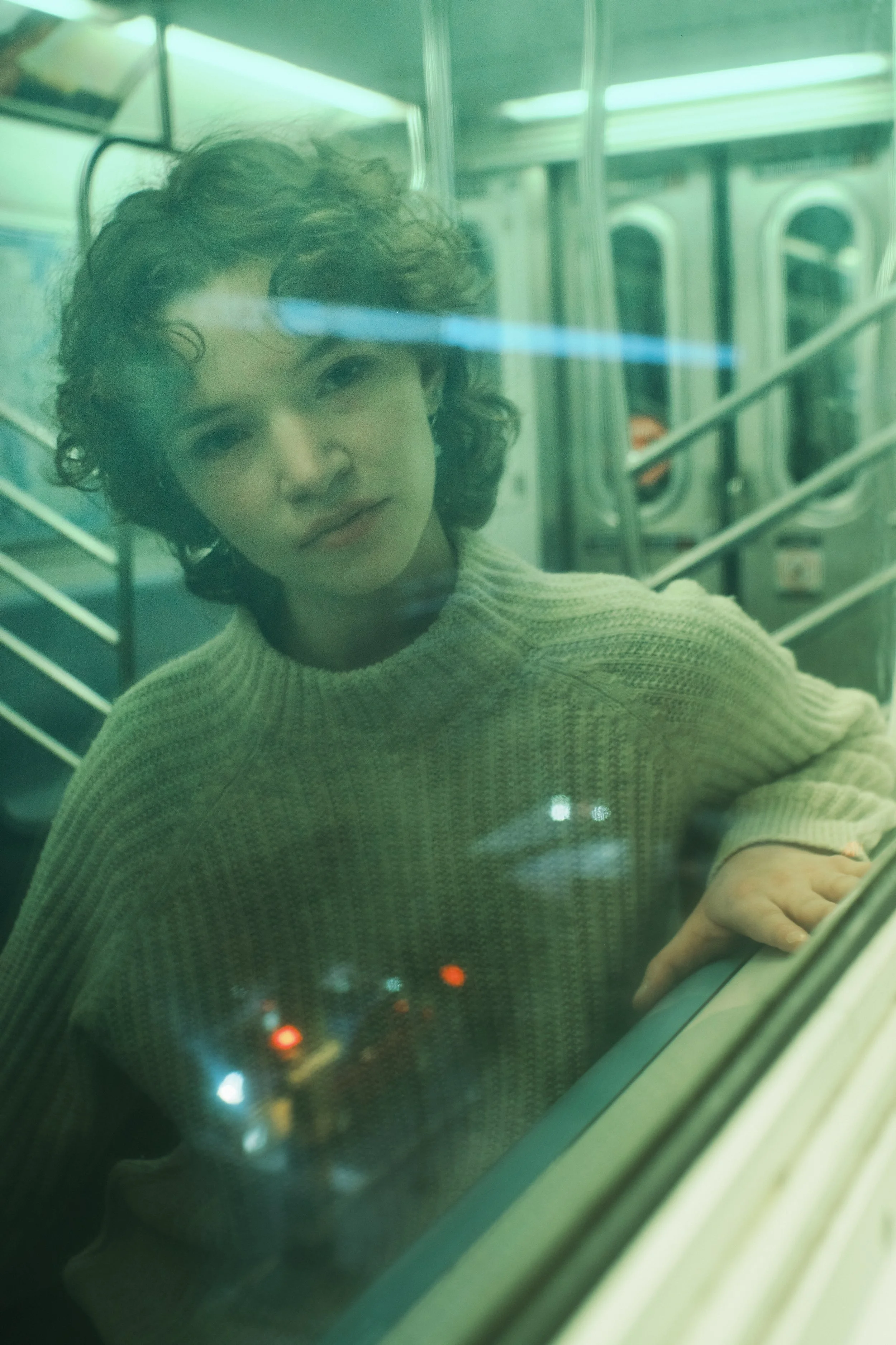 A woman with curly hair wearing a beige sweater looks through a train window at night, with city lights reflecting on the glass.  JSnaps Photography New York City Fashion lifestyle photography 