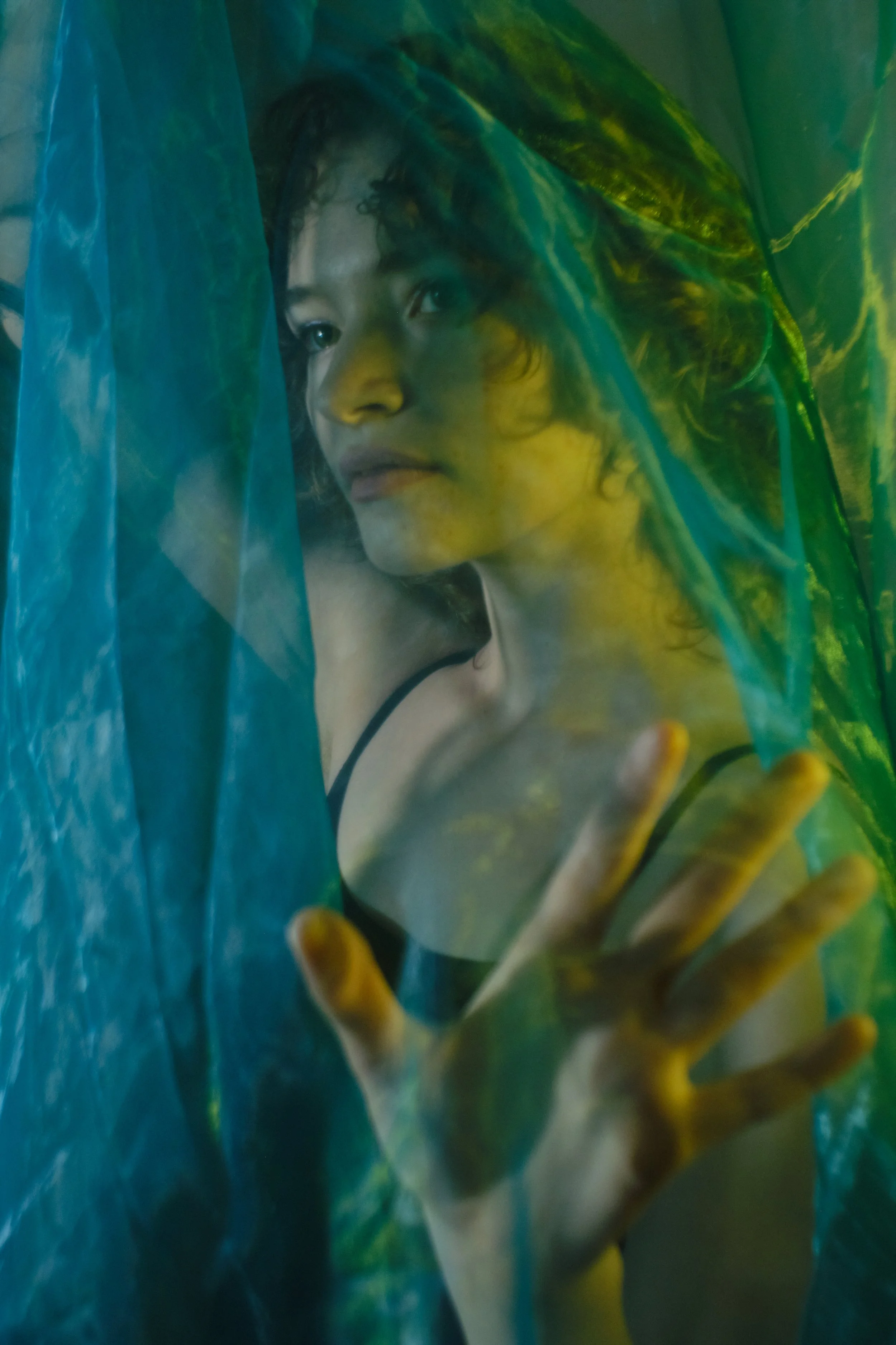 A woman with curly hair and a black strap top is behind a semi-transparent green and blue plastic sheet, making a peace sign with her hand. Portrait Photographer New York City