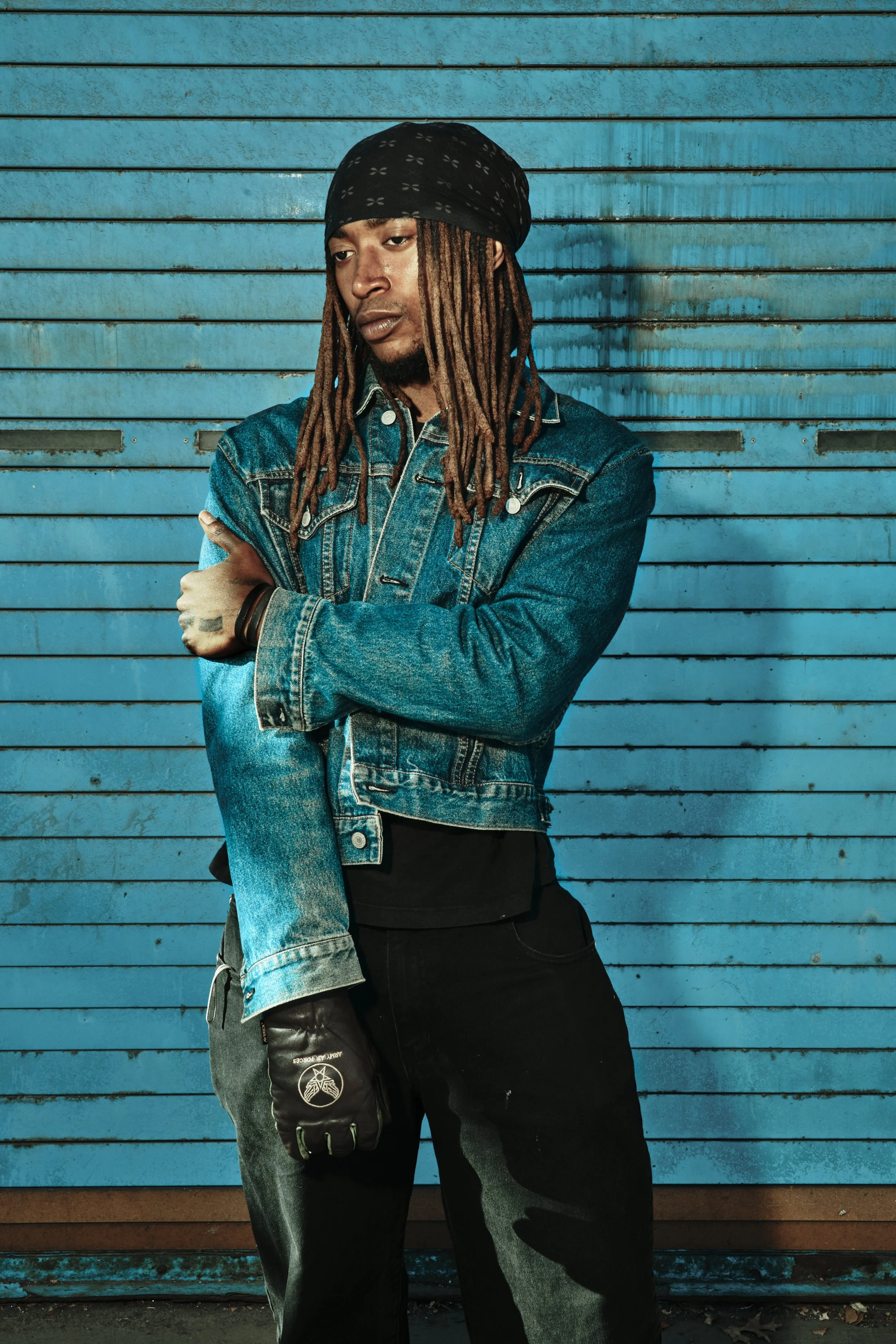 A young man with dreadlocks wearing a black bandana, denim jacket, black pants, and a black glove with a white logo, standing in front of a blue, weathered, metal roll-up door. JSnaps Photography New York City Fashion lifestyle photography 