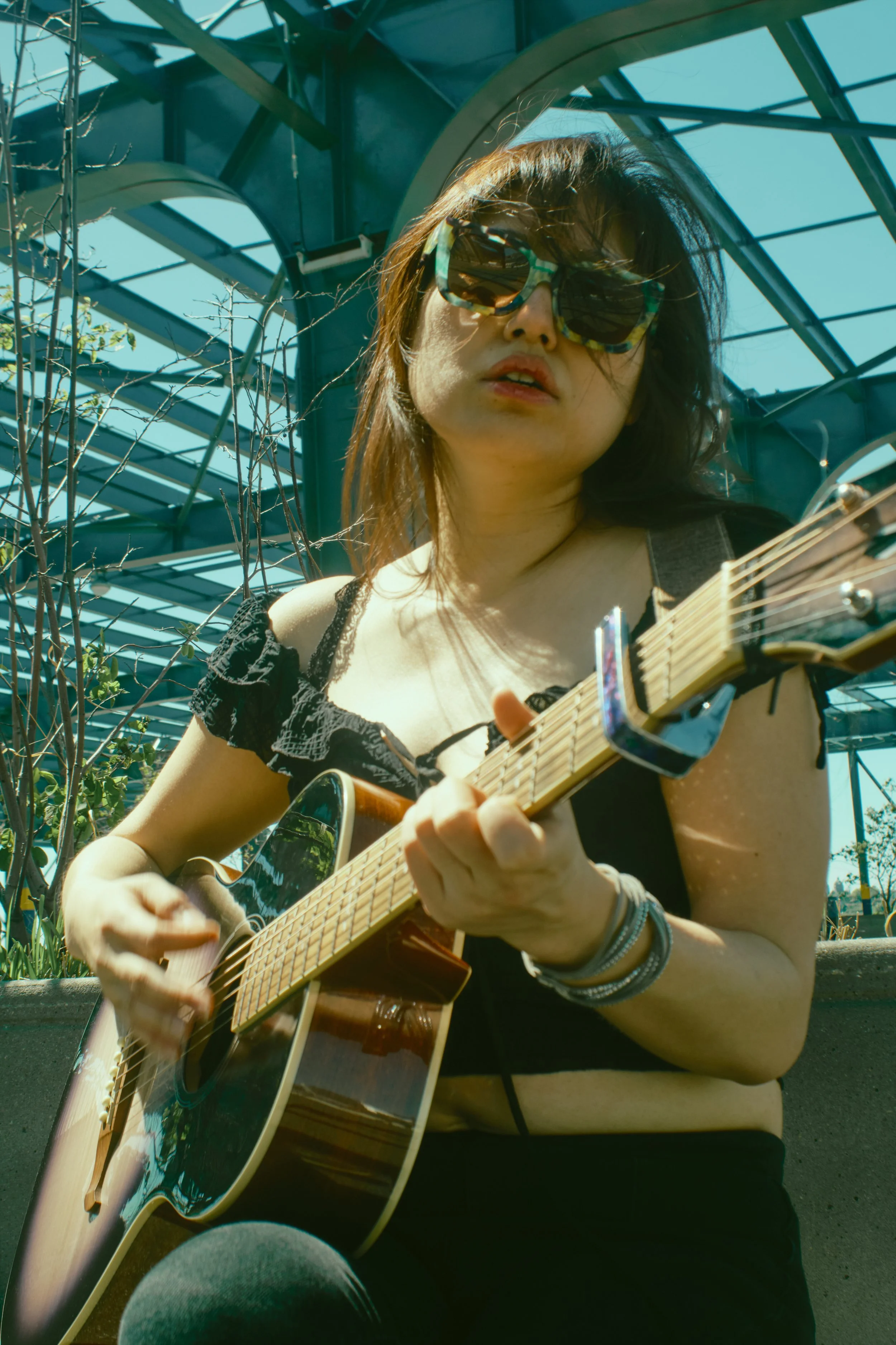 A woman with sunglasses playing guitar outdoors against a backdrop of a glass greenhouse and blue sky. JSnaps Photography New York City Fashion lifestyle photography 